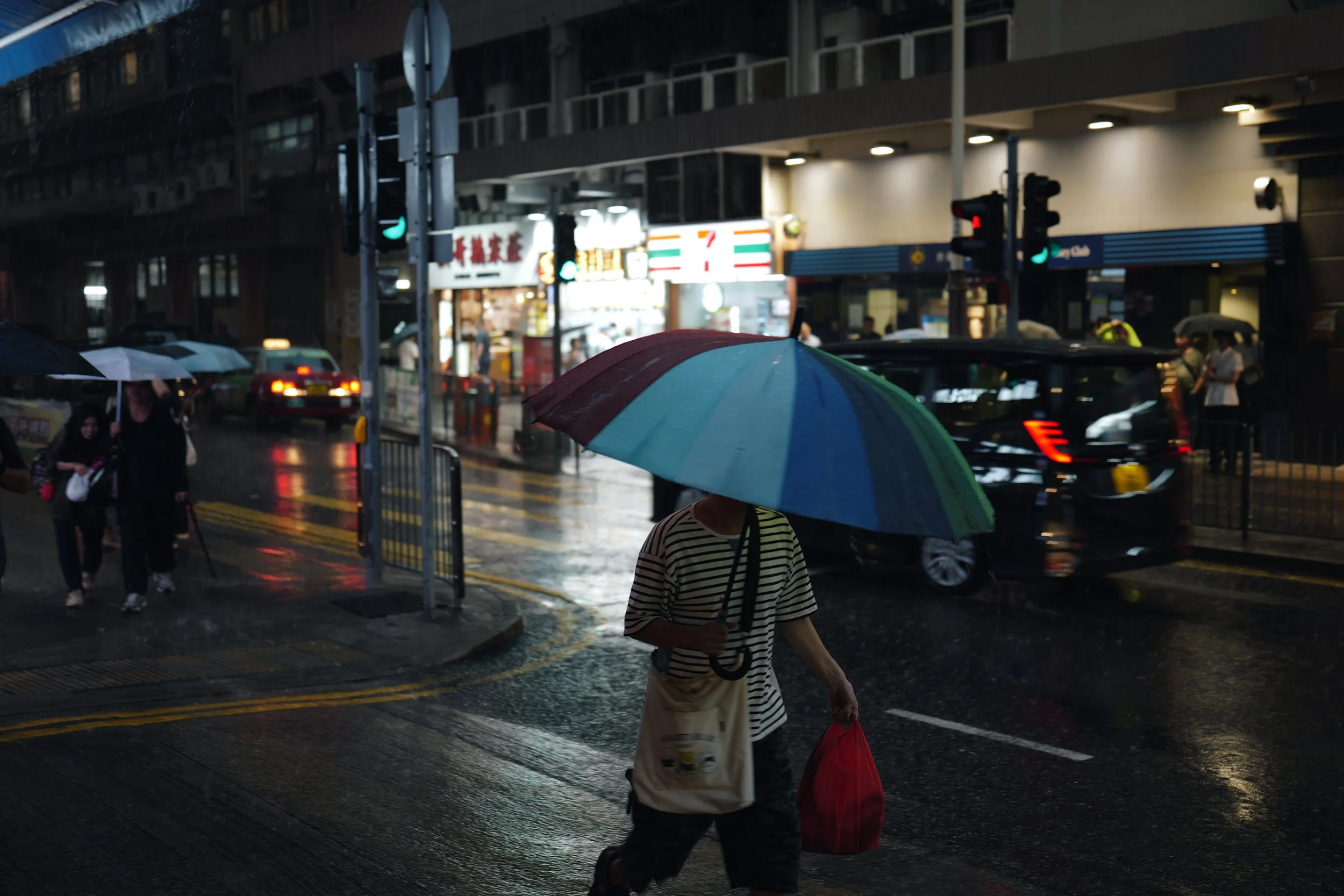 People walking with umbrellas in the rain on a city street at night, with cars and illuminated storefronts visible.