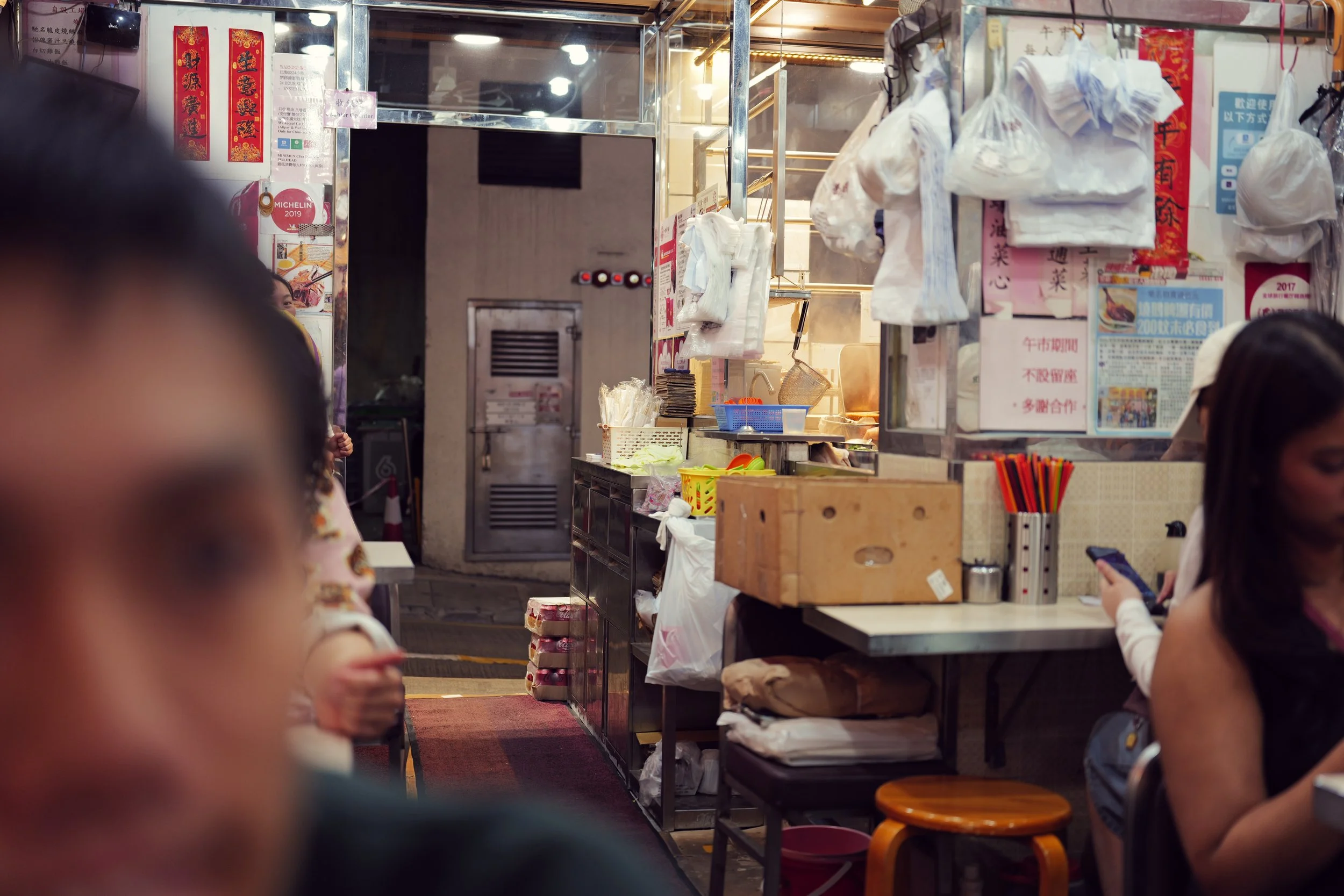 Inside a small Asian restaurant or food stand with hanging white plastic bags and colorful posters on the wall, people sitting and waiting, with some children near the counter.