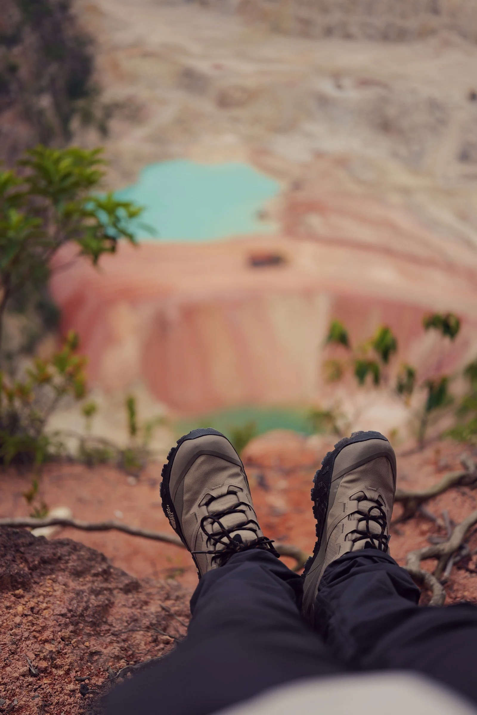Hiking boots extend over the red-earth overlook above the blurred turquoise pool.