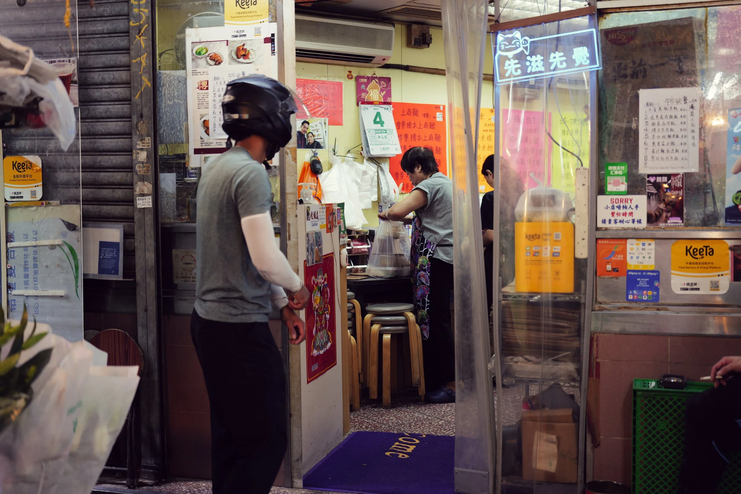 Inside a small Asian restaurant or food stall, a man with a helmet is in the foreground, and a woman in an apron is preparing food behind the counter. The interior features colorful signs and posters, some with Chinese characters, and various food an