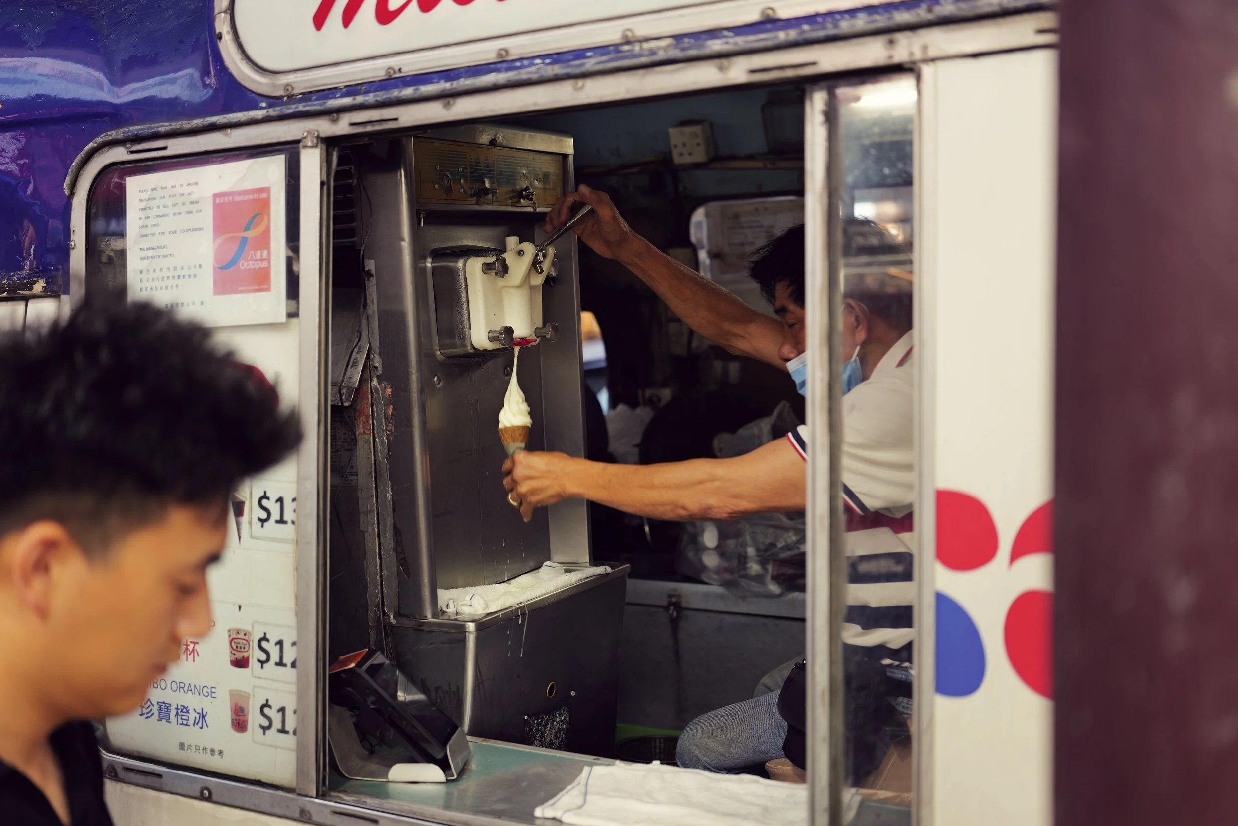 A person in a white shirt and face mask serving ice cream from a soft serve machine at a food stand, with part of another person in the foreground and a poster with drink options in Asian characters and prices visible on the stand.