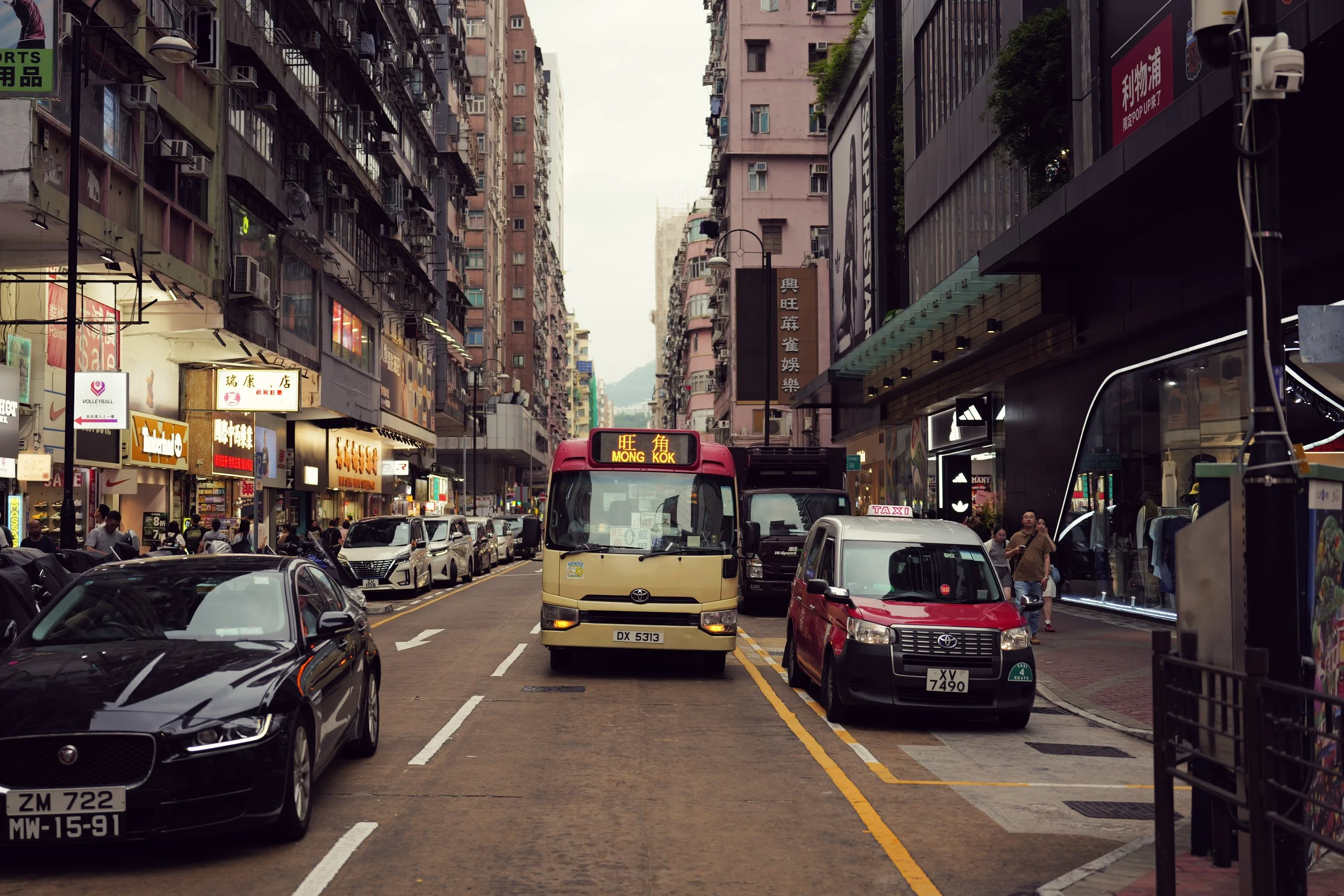 City street scene with cars, buses, and pedestrians, surrounded by tall buildings with commercial signs and storefronts.