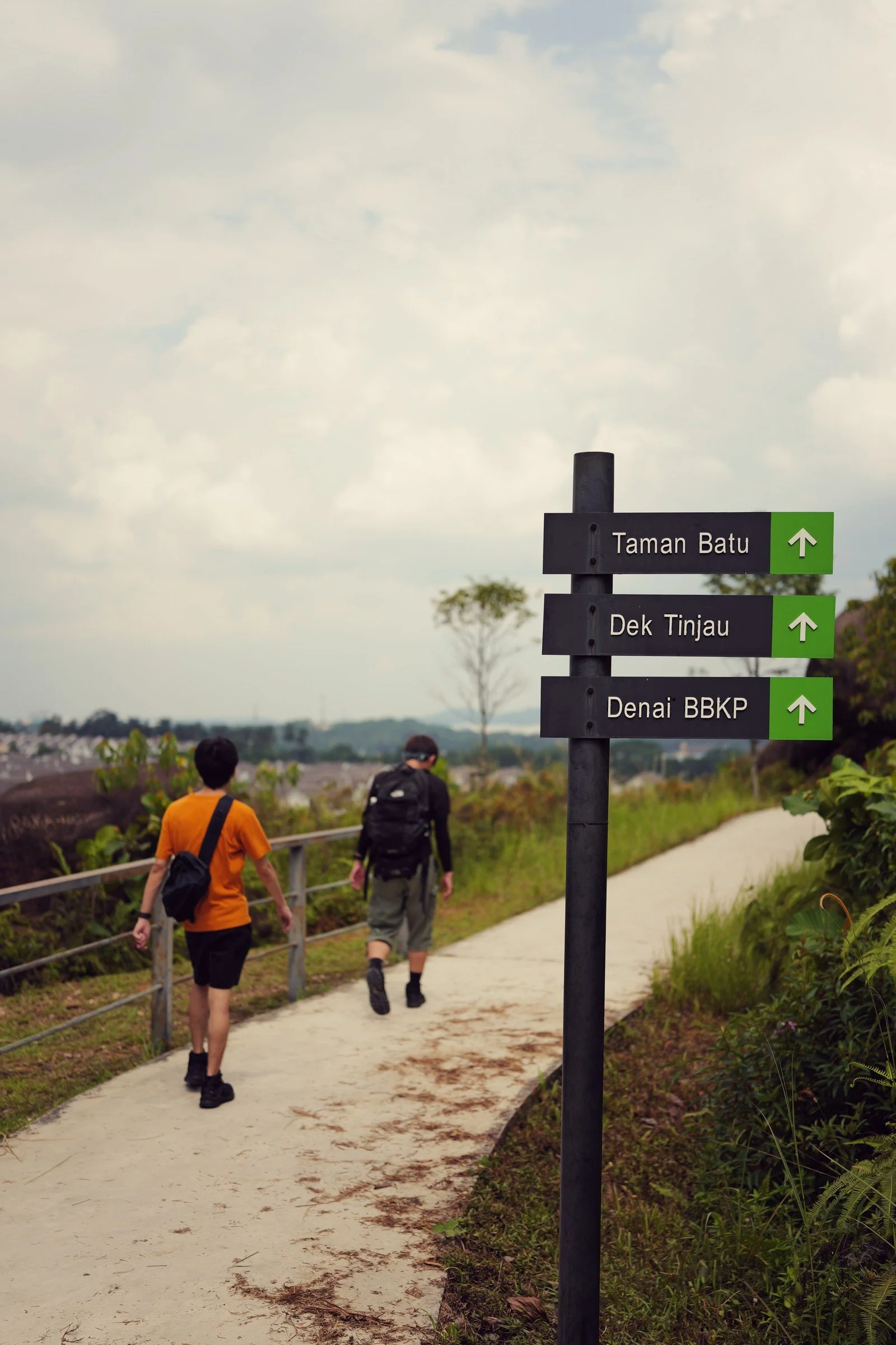 Two hikers walk past a trail sign beneath a cloudy sky.