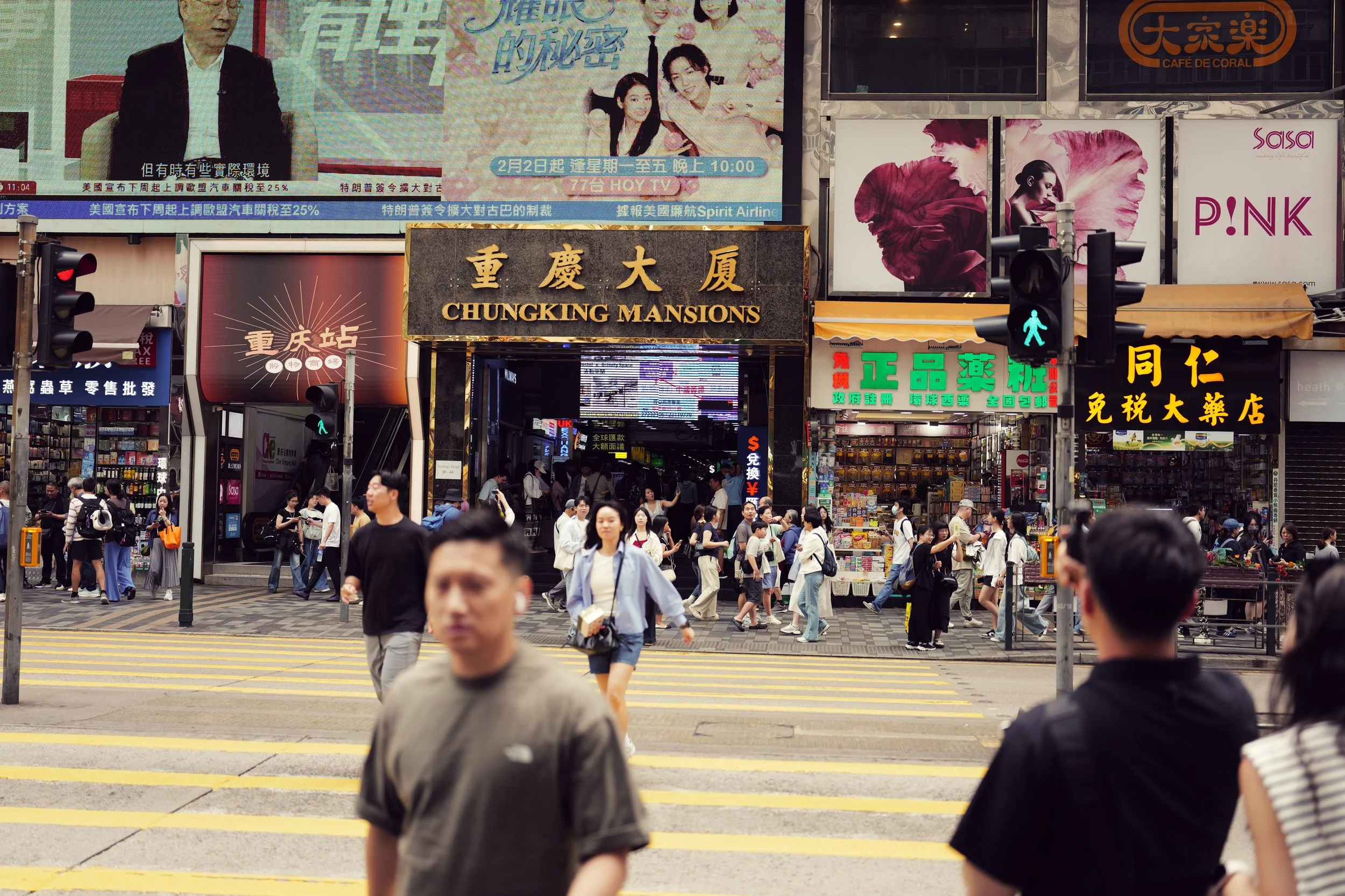 A busy city street scene with numerous pedestrians crossing at a crosswalk, numerous advertising billboards and shop signs above the sidewalk, including a sign for Chungking Mansions. The crowd includes men and women of various ages, some wearing mas