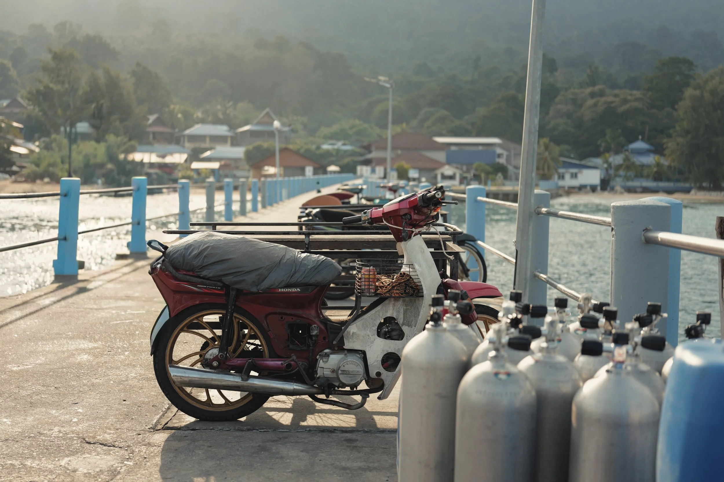 A red motorbike parked on a dock near the water, with propane tanks beside it and houses on a hill in the background.
