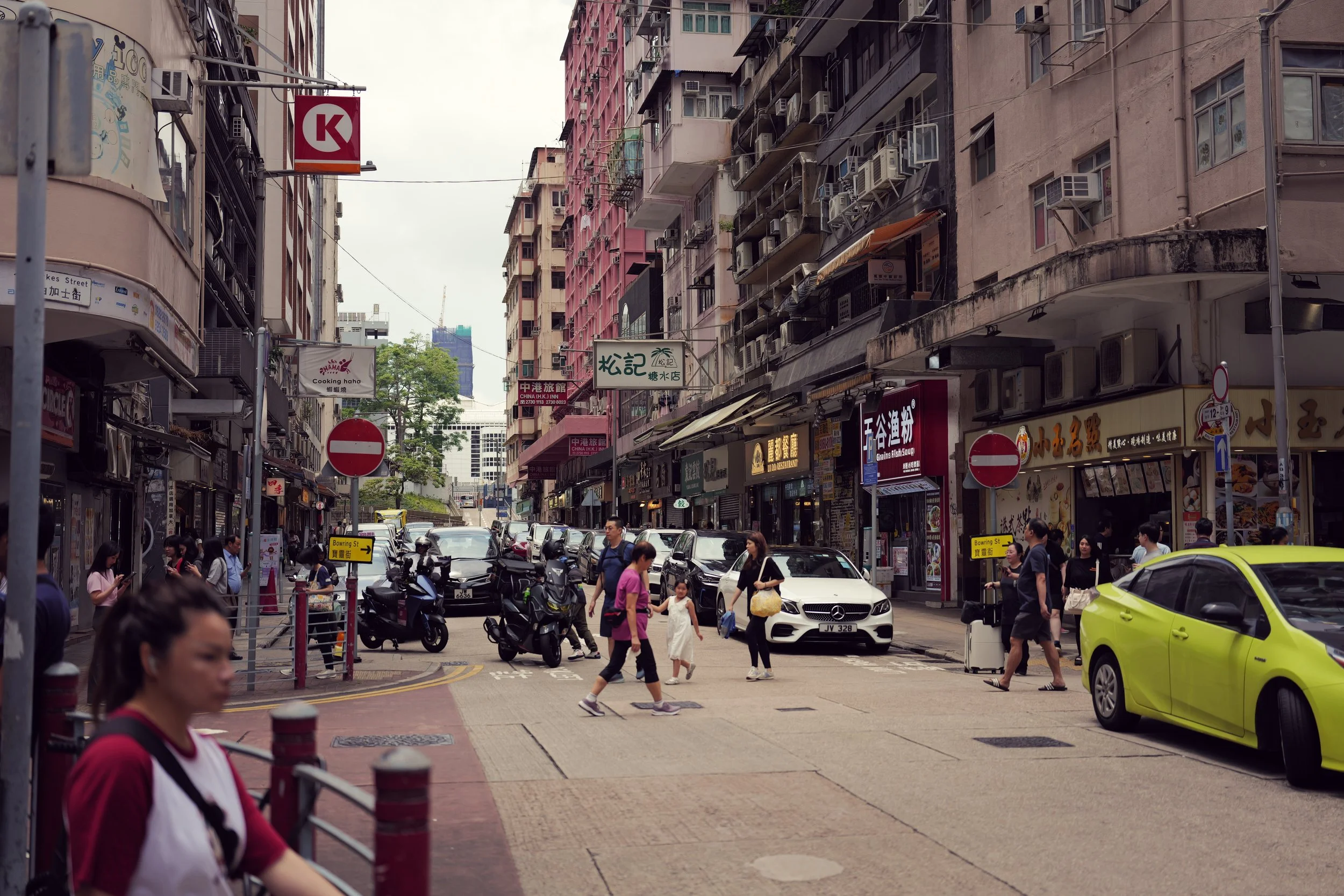 City street scene with pedestrians crossing, parked and moving cars, tall buildings with commercial signs, and numerous air conditioning units on building exteriors.