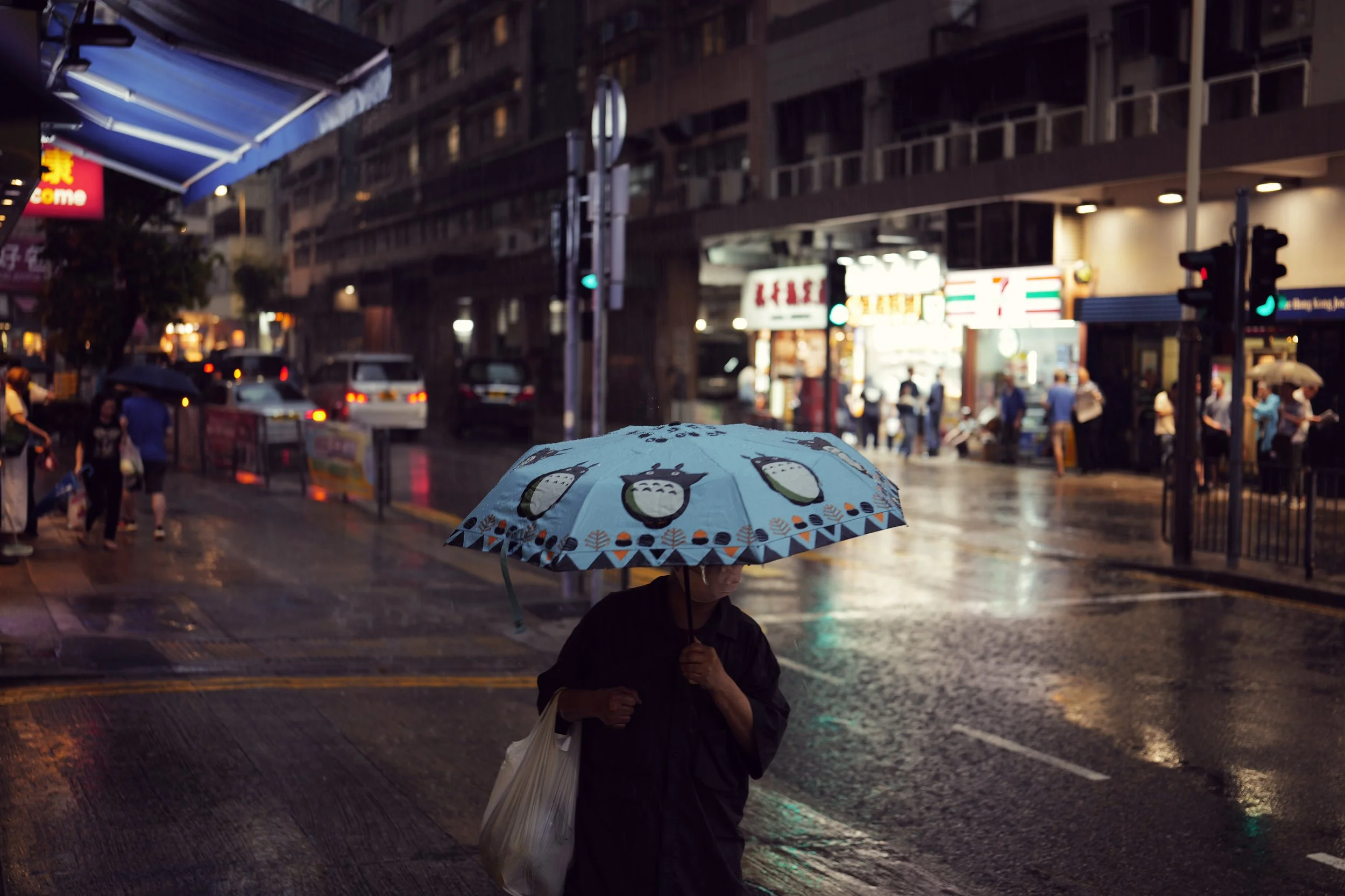 A person holding a patterned umbrella walking in the rain on a city street at night, with cars and pedestrians visible in the background.