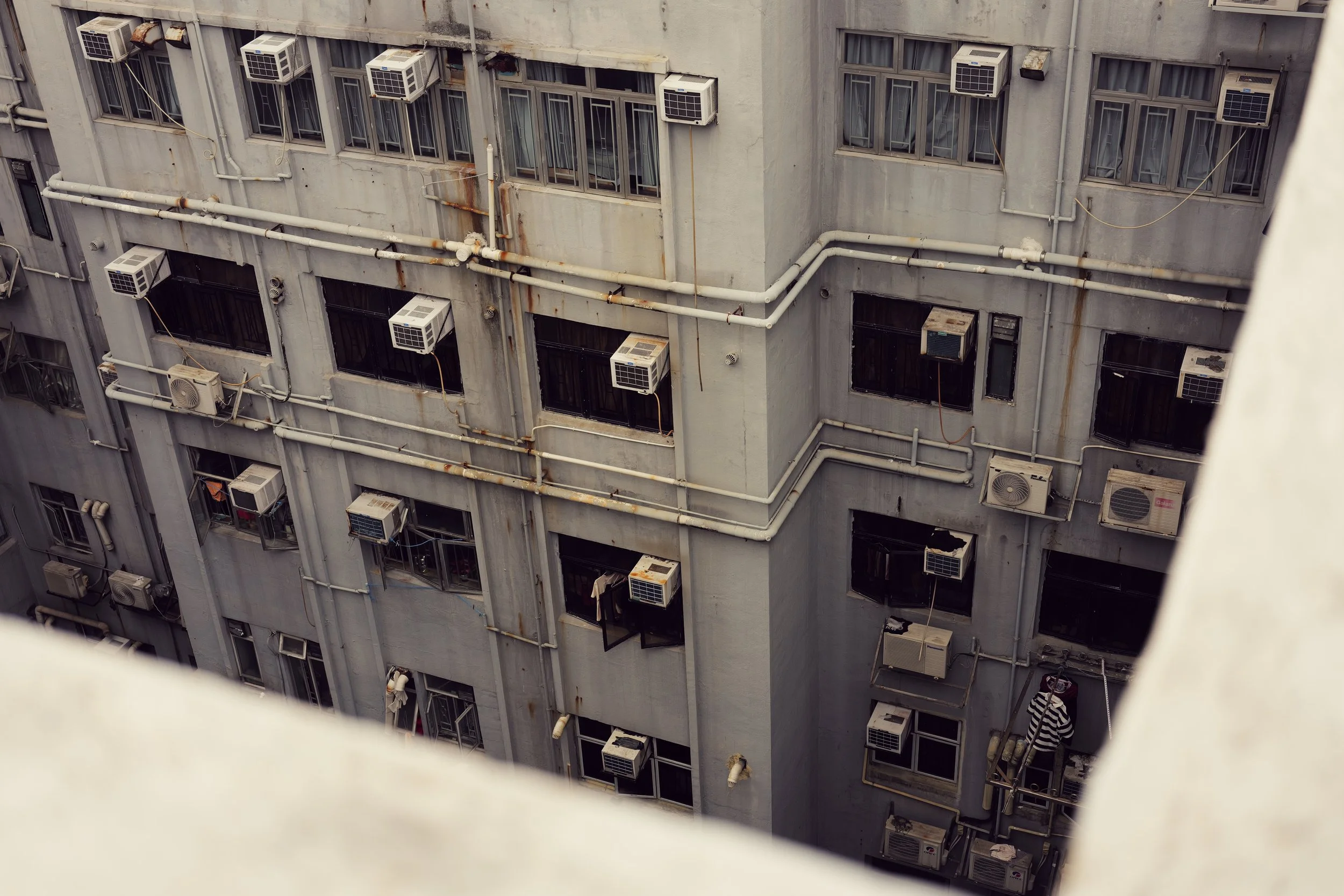 View of an apartment building's exterior with numerous windows and air conditioning units, some with rust and rust stains, and visible pipes running across the facade.