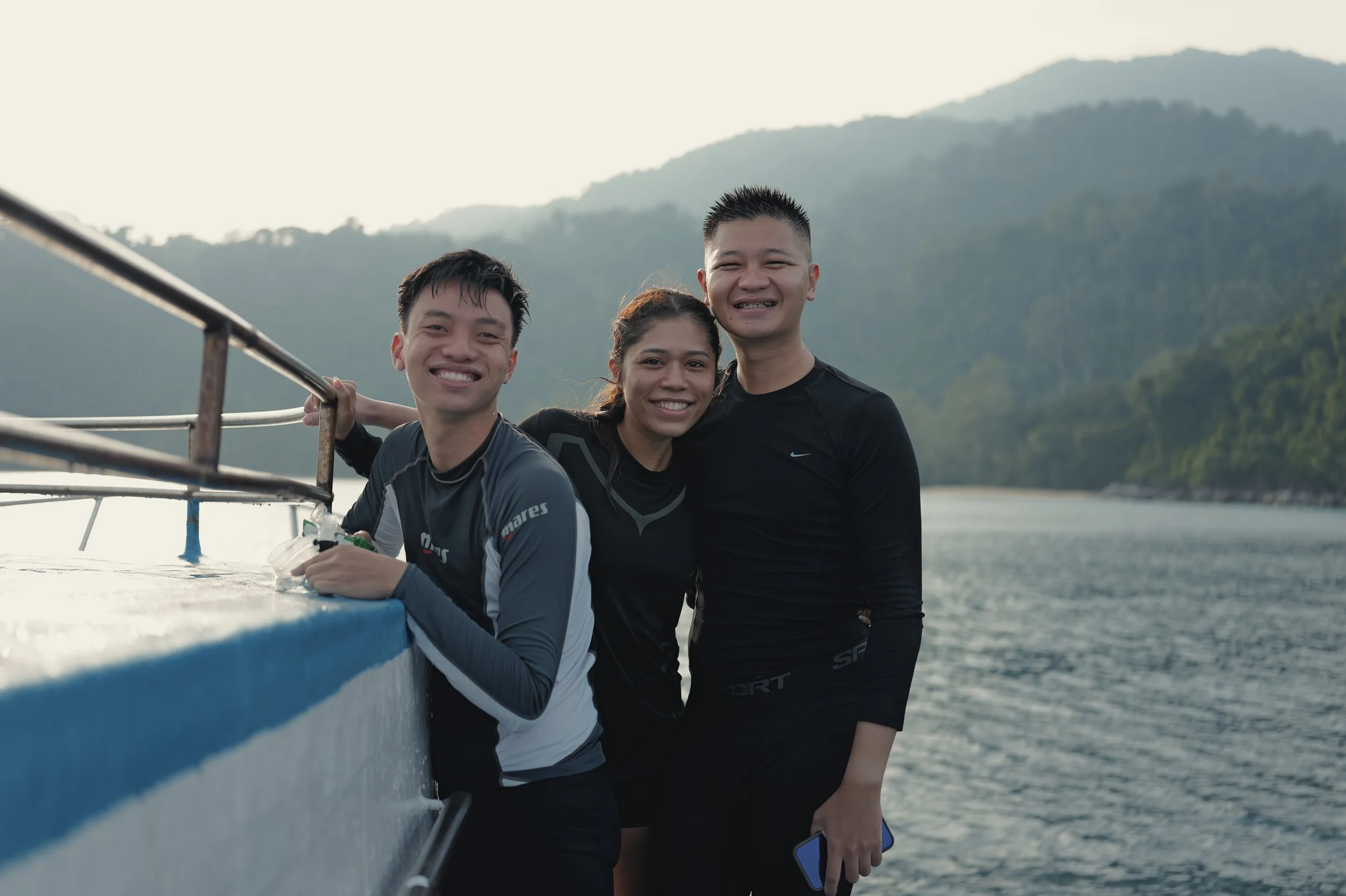 Three smiling people, two men and one woman, standing on a boat with a scenic mountainous background during daytime.