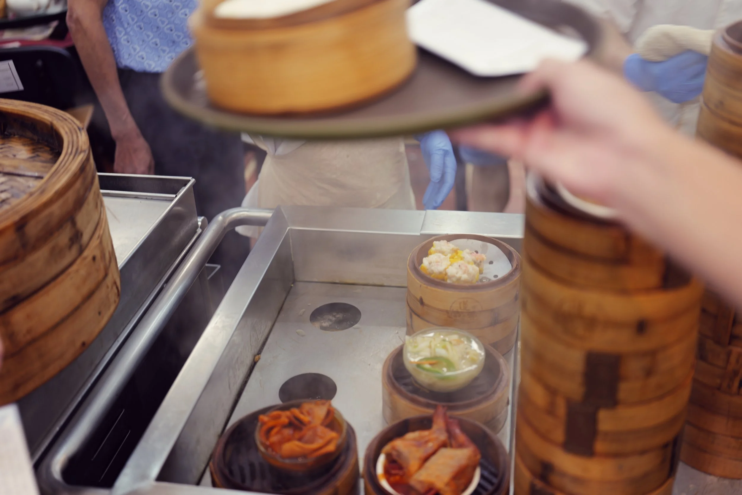 A person serving food at an Asian buffet with round bamboo steamers and small cups of dim sum on a metal counter.
