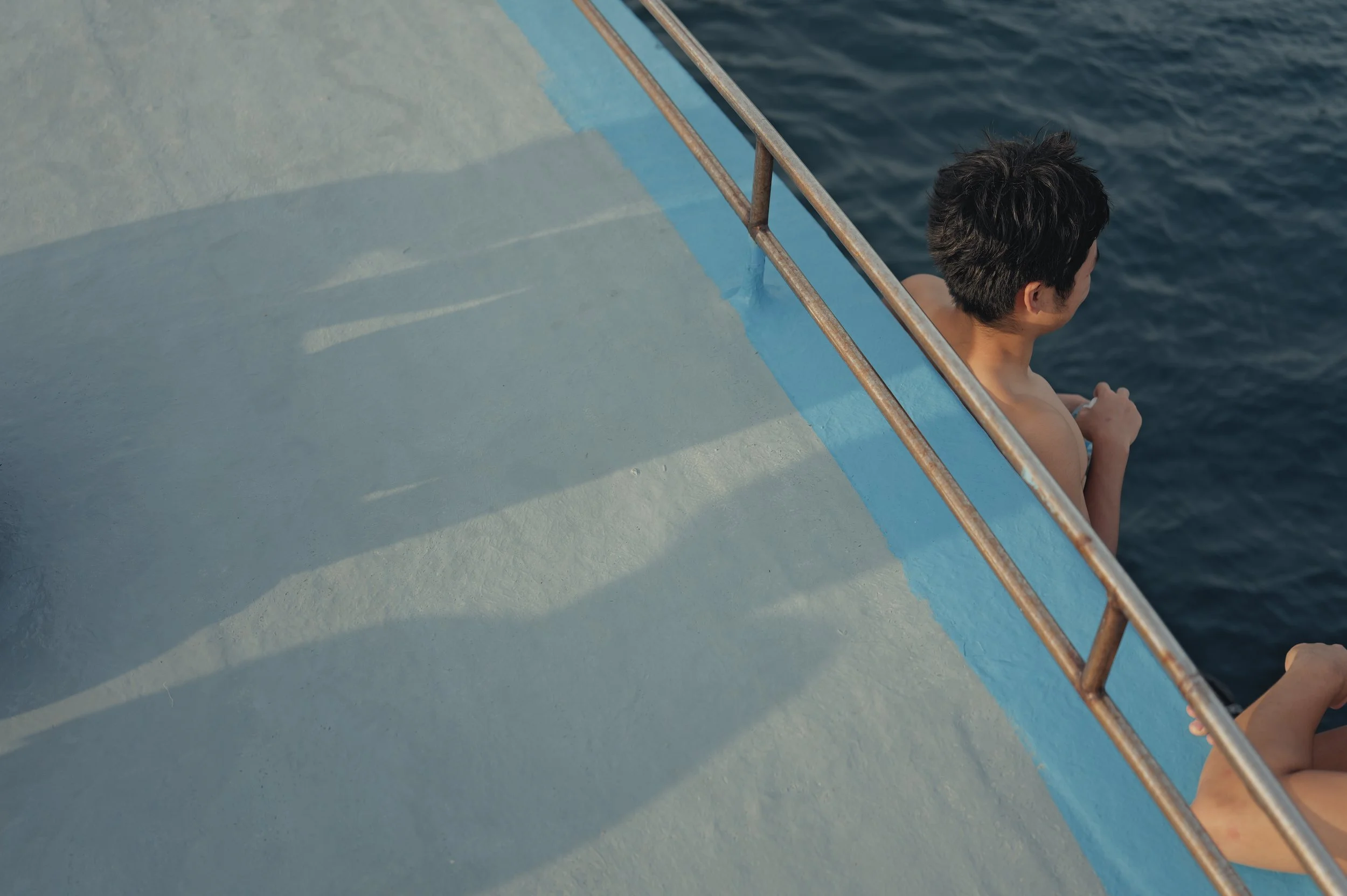A person with short dark hair sitting on a boat or dock, looking out over the water, with their back to the camera, surrounded by a metal railing and blue-painted surface.
