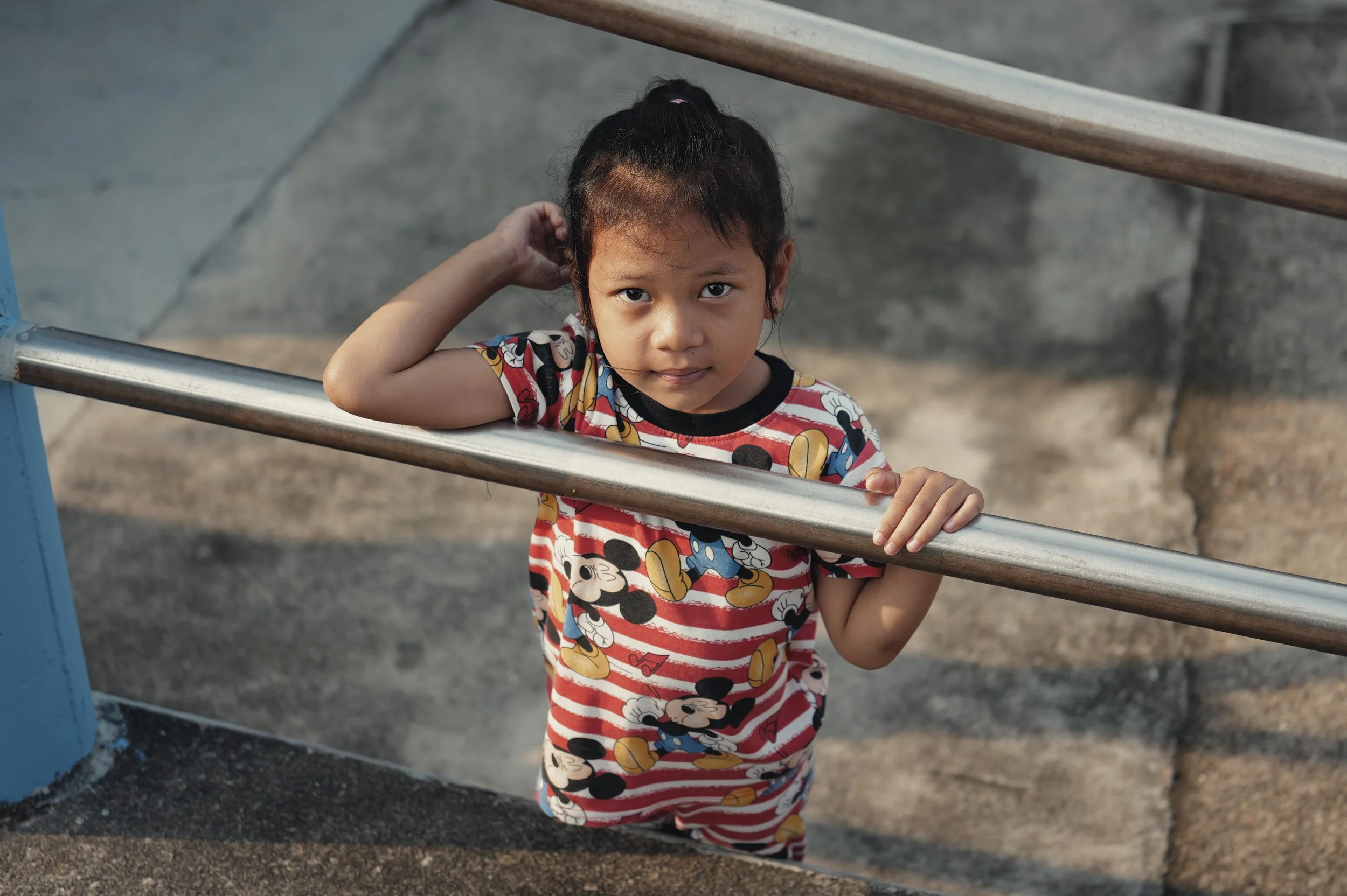 A young girl with dark hair and light brown skin, wearing a red and white striped Mickey Mouse t-shirt and matching shorts, stands outdoors at a playground. She has her left hand behind her head and is holding onto a metal bar with her right hand, lo
