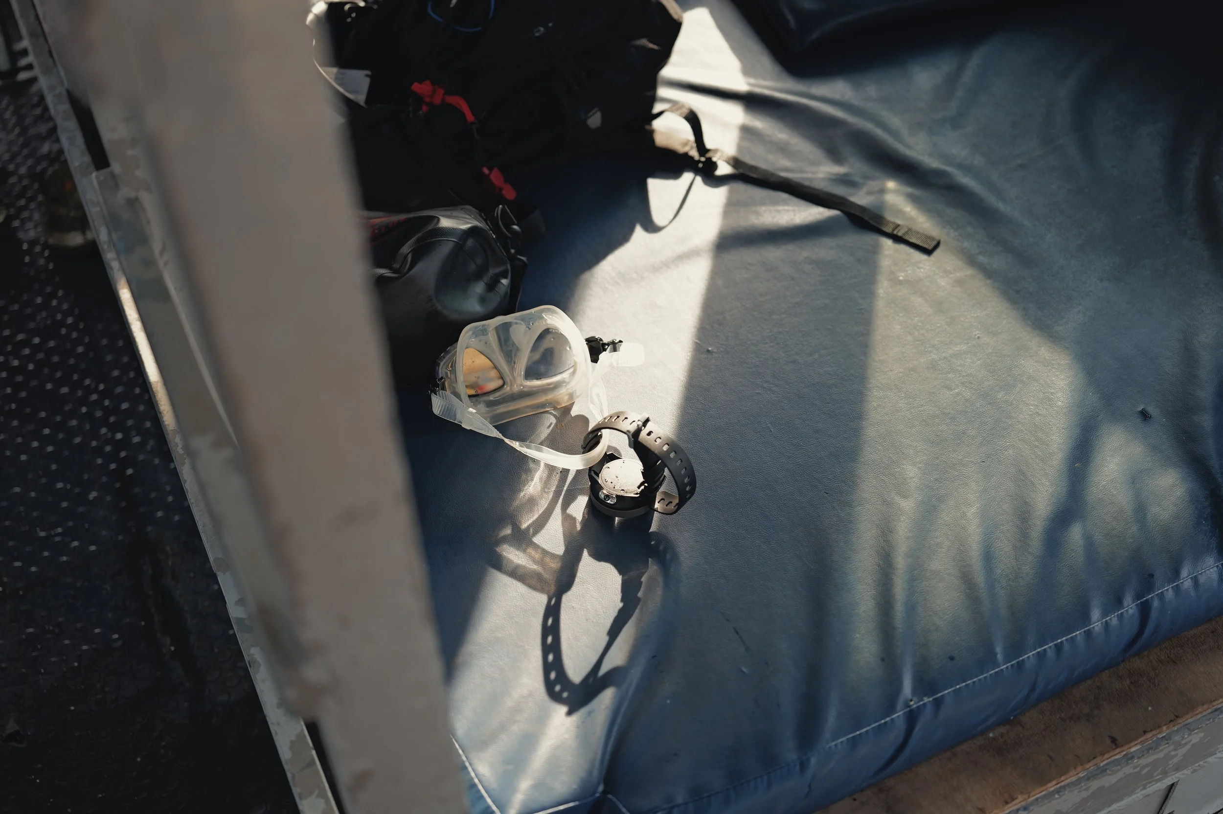 A swimming mask with a snorkel and a wristwatch placed on a silver metallic boat surface, with backpacks visible in the background.