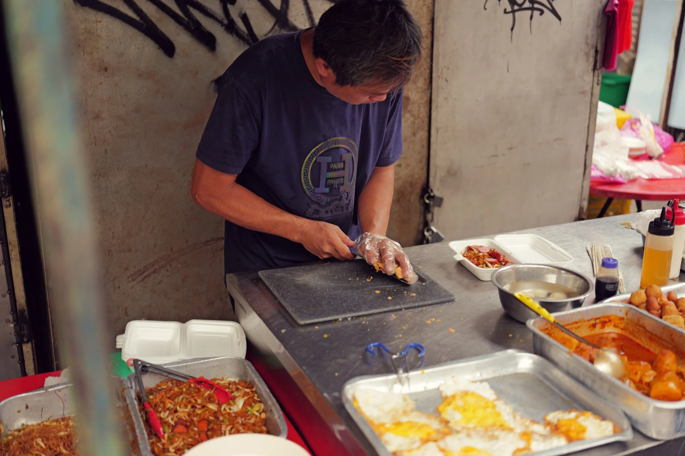 A cook slices food at a metal stall counter surrounded by trays of ingredients.