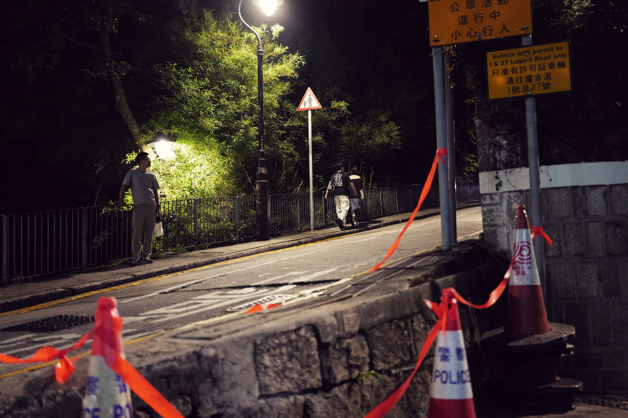 Night scene of a sidewalk with orange caution cones and police tape in the foreground, a street lamp illuminating trees and pedestrians walking along the sidewalk, and multiple signs including a prone pedestrian warning and traffic information in Asi