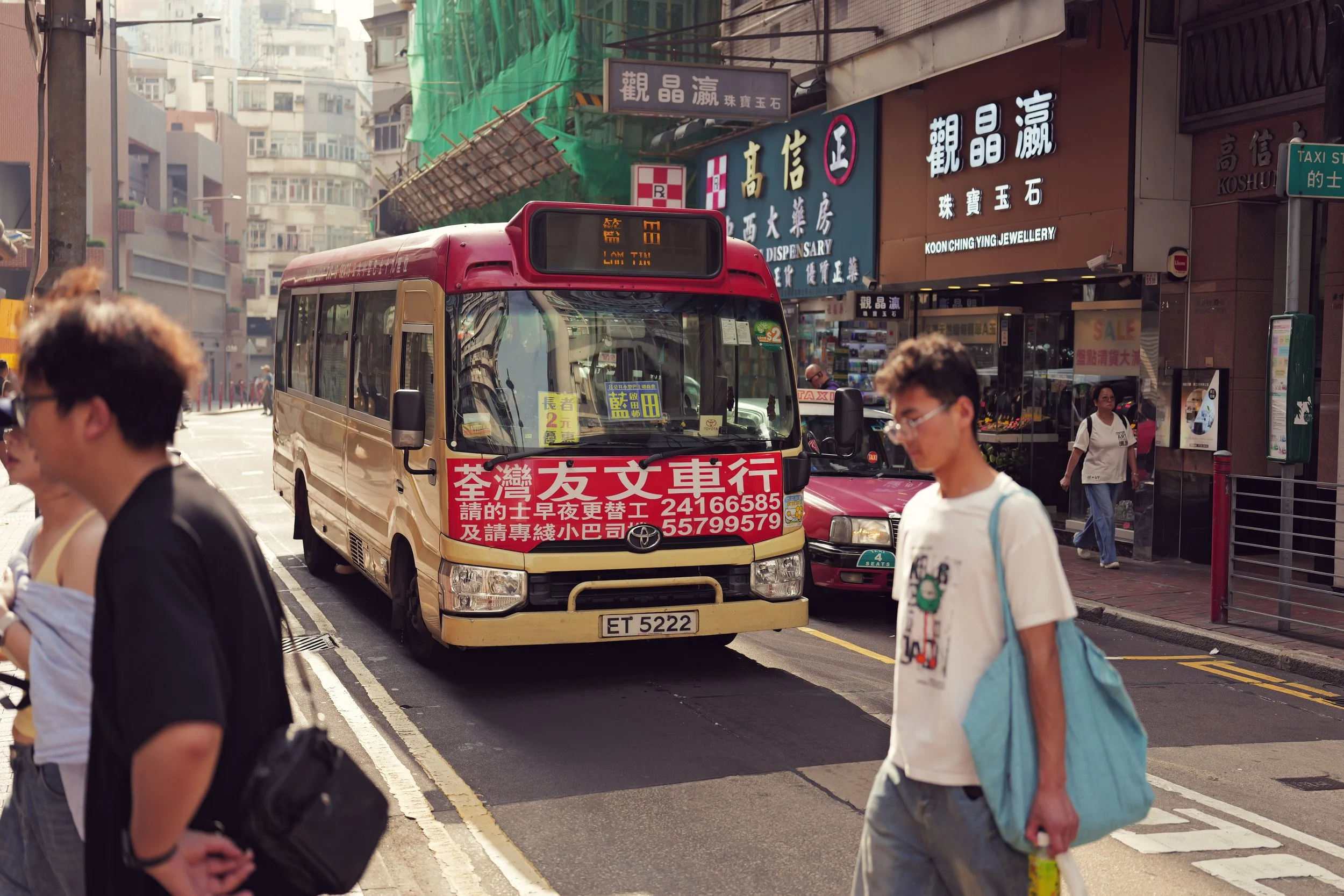 A busy city street scene with pedestrians crossing the road and a small bus in the foreground. The bus has signage in Chinese characters and a license plate number. Commercial signs in Chinese and English are visible on buildings, indicating shops an