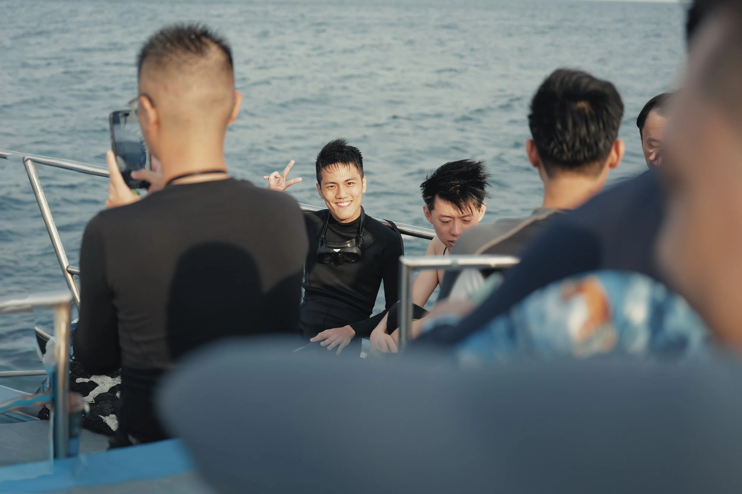 Group of people on a boat near the water, with one person making a peace sign and smiling, while others are looking at the water or camera, some taking photos.