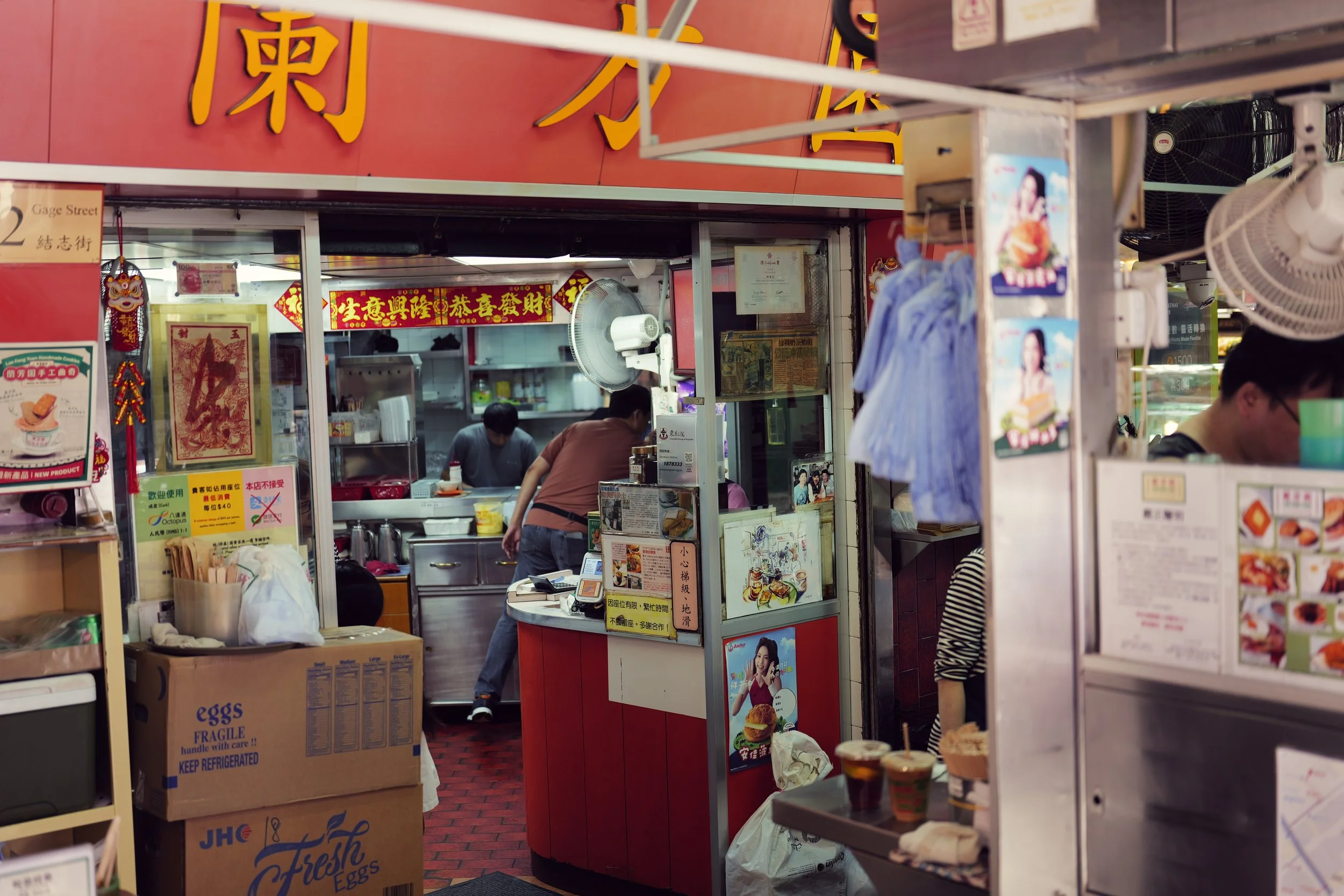 A small food stall or shop with red signage, Asian decorations, and posters. There are a few people working inside, and various signs and advertisements on display. A cardboard box labeled "Eggs" is visible in the foreground.