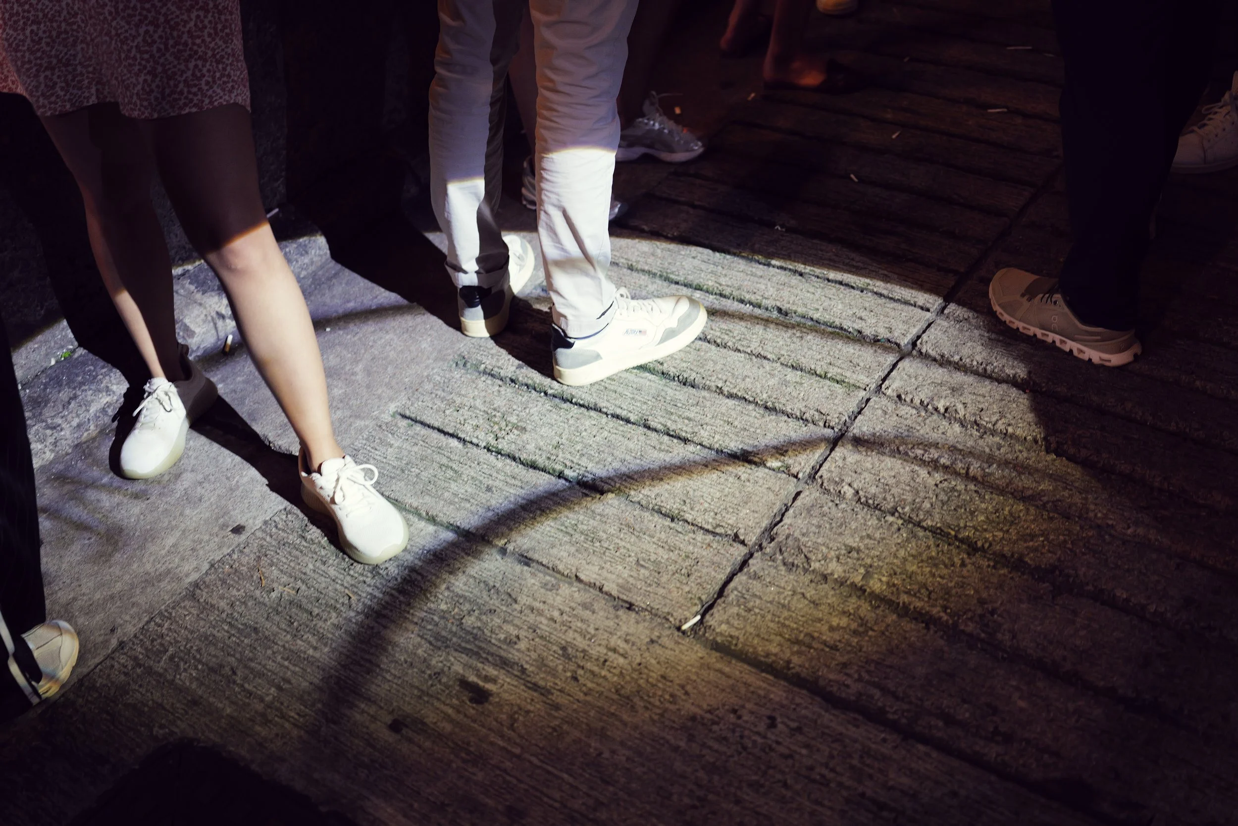 People standing on a wooden deck and concrete sidewalk, wearing sneakers and casual clothing, during the evening or night.