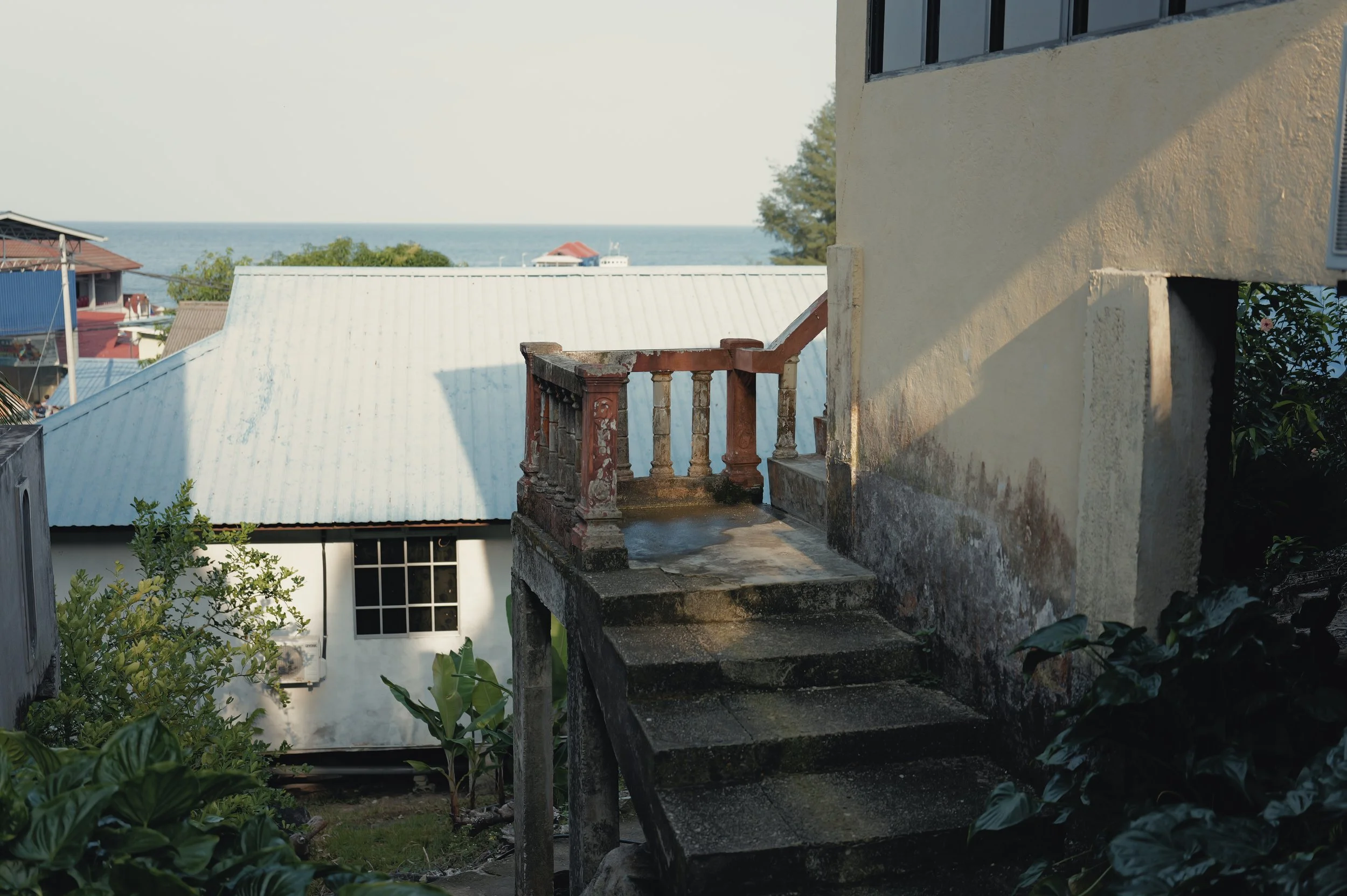 View of rooftops with metal and tiled roofs near the ocean, with a yellow building and a small balcony in the foreground, and trees and ocean in the background.