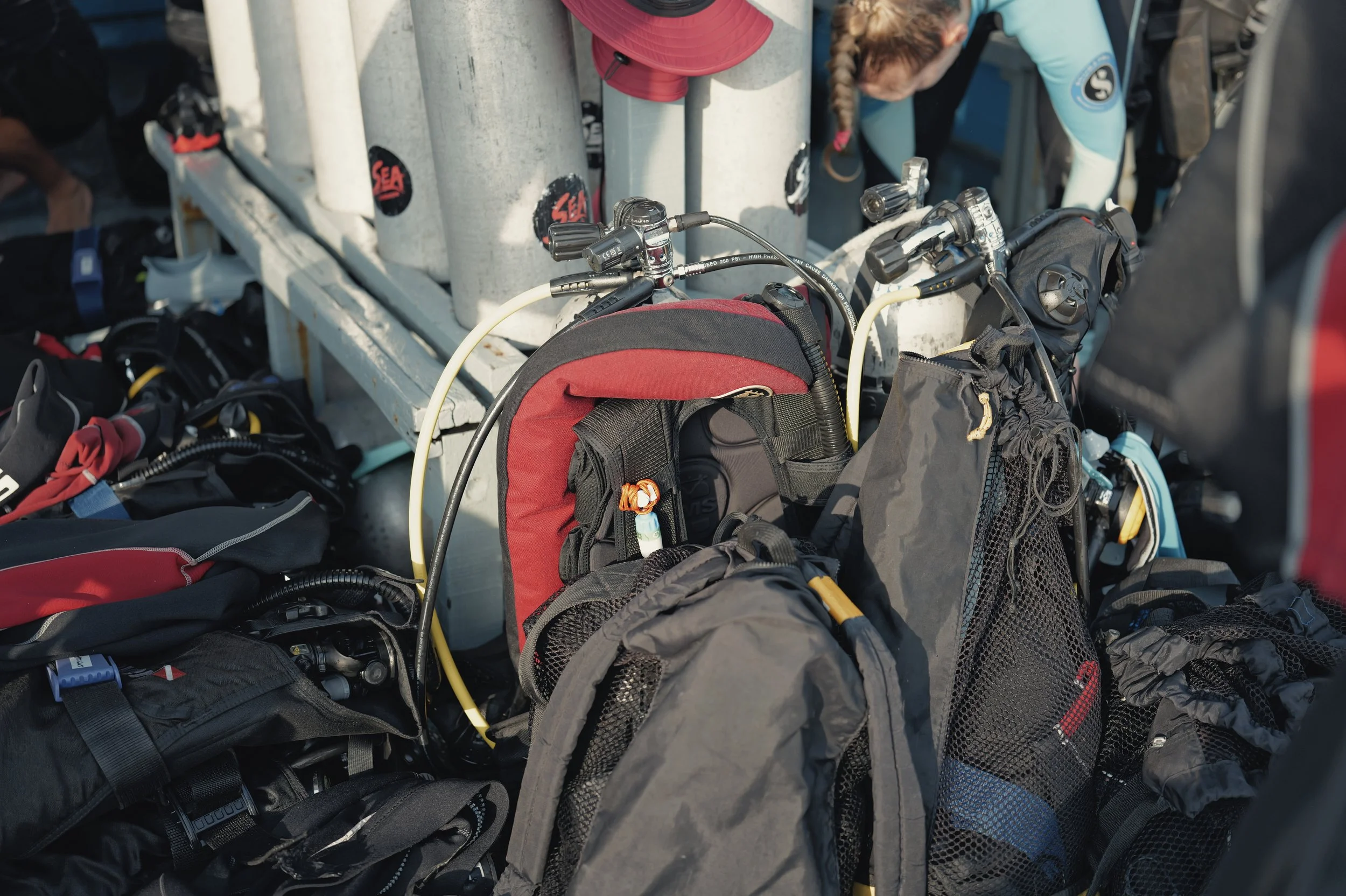 Close-up of scuba diving gear, including tanks, regulator, and various bags and equipment, with a person wearing a wetsuit working in the background.