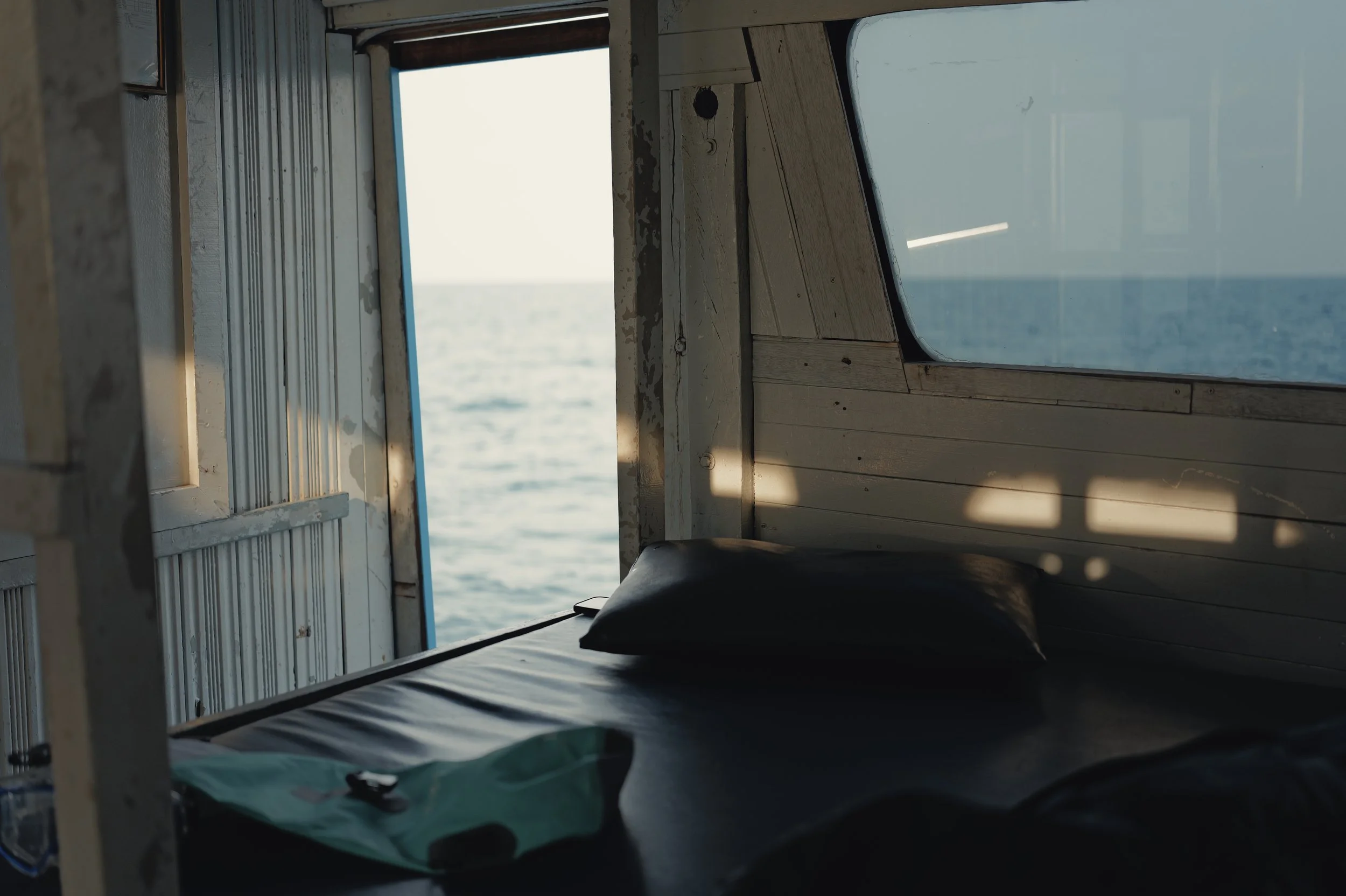 The image shows the inside of a boat or camper, with a bed and a window overlooking the water. The interior has wooden paneling and a pillow on the bed. Shadows from the boat's structure create patterns on the bed. The scene outside the window reveal