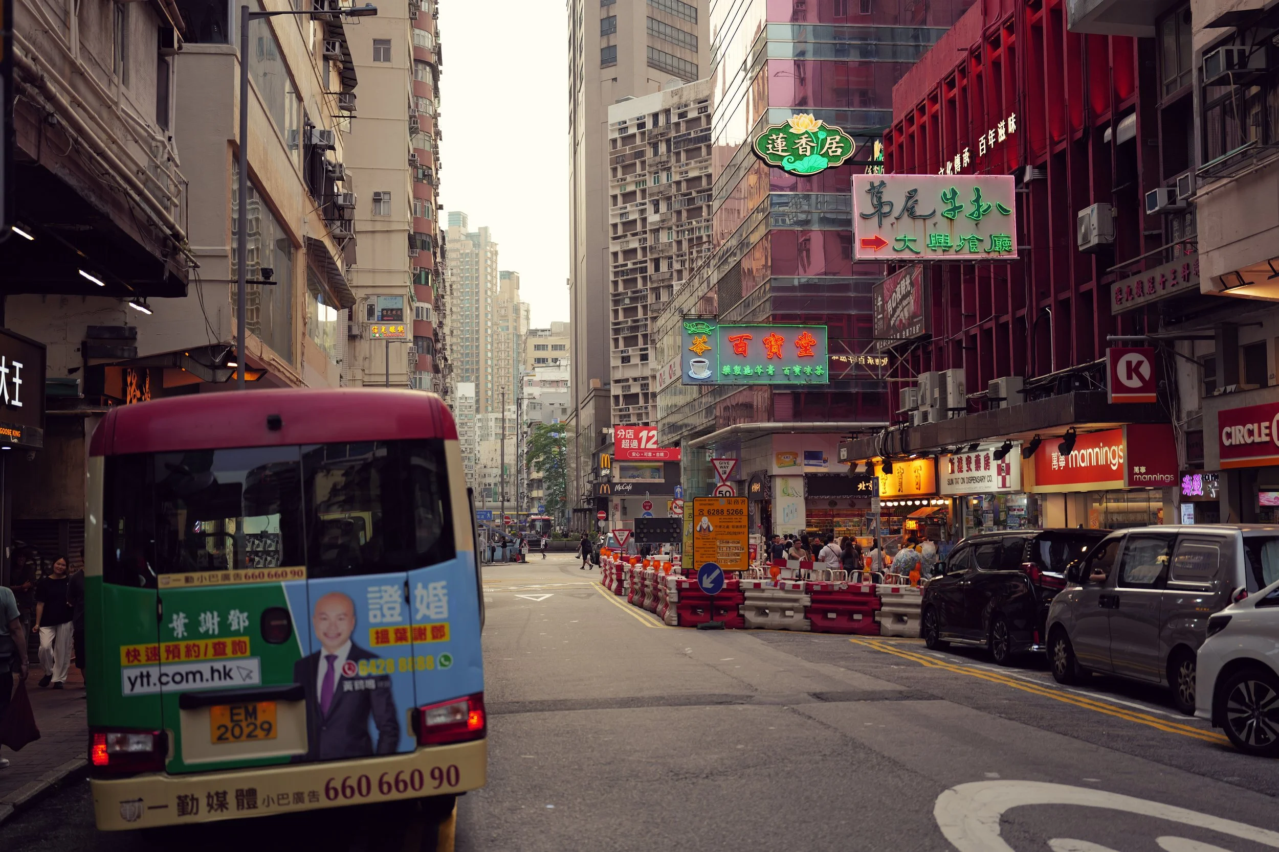 A busy city street scene with tall buildings, neon signs in Chinese, a colorful bus in the foreground, and pedestrians walking on the sidewalks.