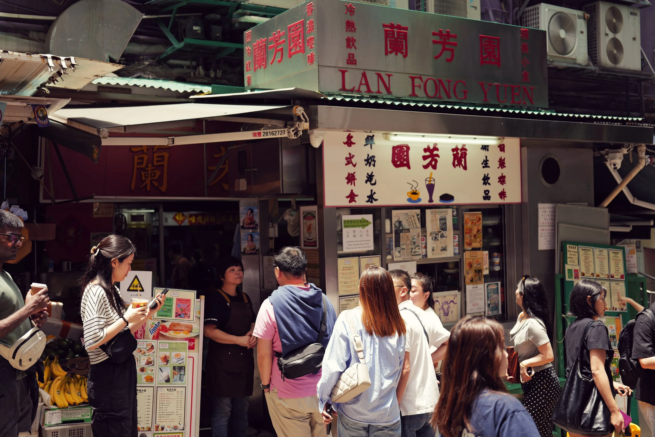Crowd of people standing outside a street food stall with signs in Chinese characters and English, selling drinks, snacks, and desserts.
