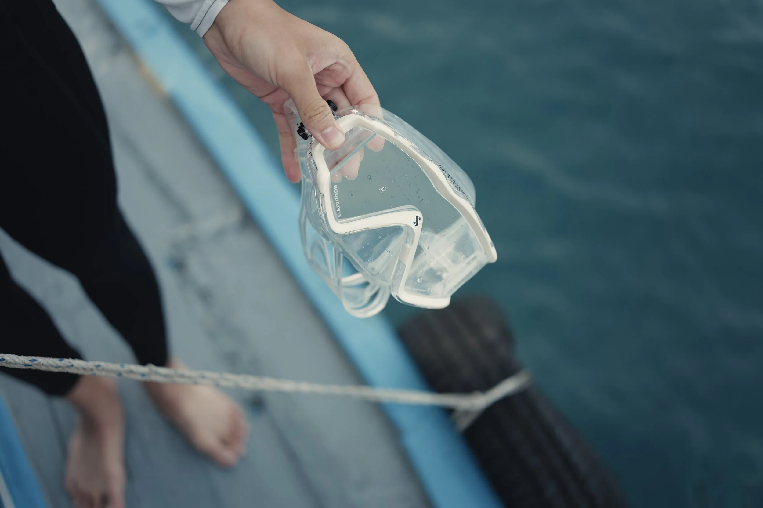 Close-up of a person's hand holding a clear snorkel mask near a body of water, with the person's feet visible on a boat or dock.