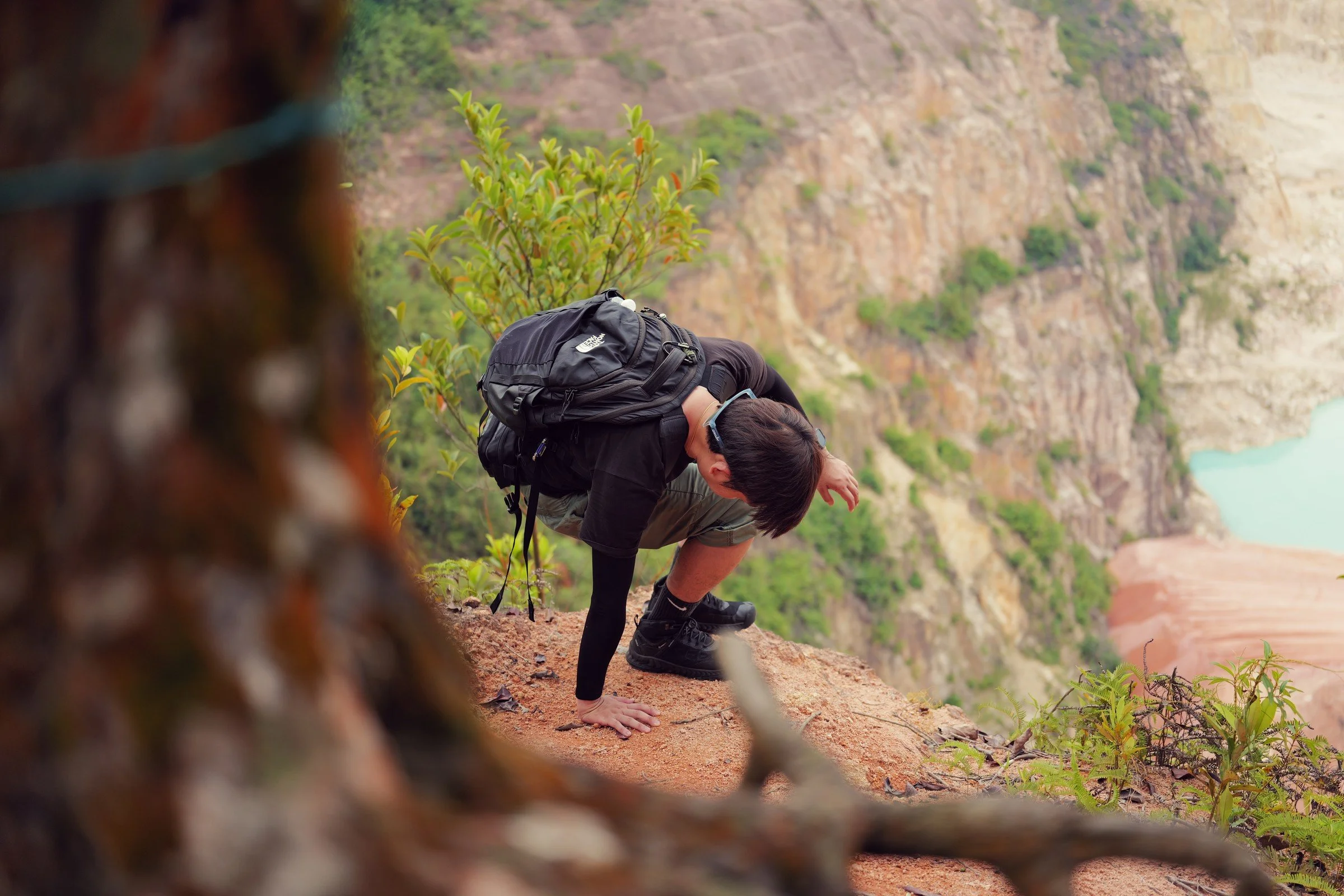 A backpacked hiker climbs a red-earth slope between trees above the lake.