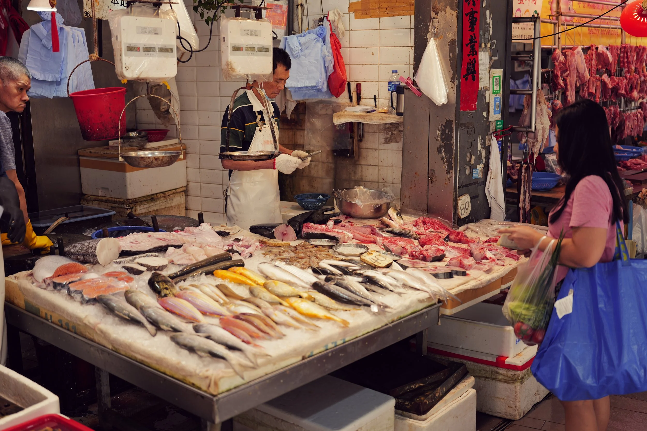 A bustling fish and meat market with vendors and customers. Displays of fish and meat on a table, some hanging, along with scales and other market supplies.