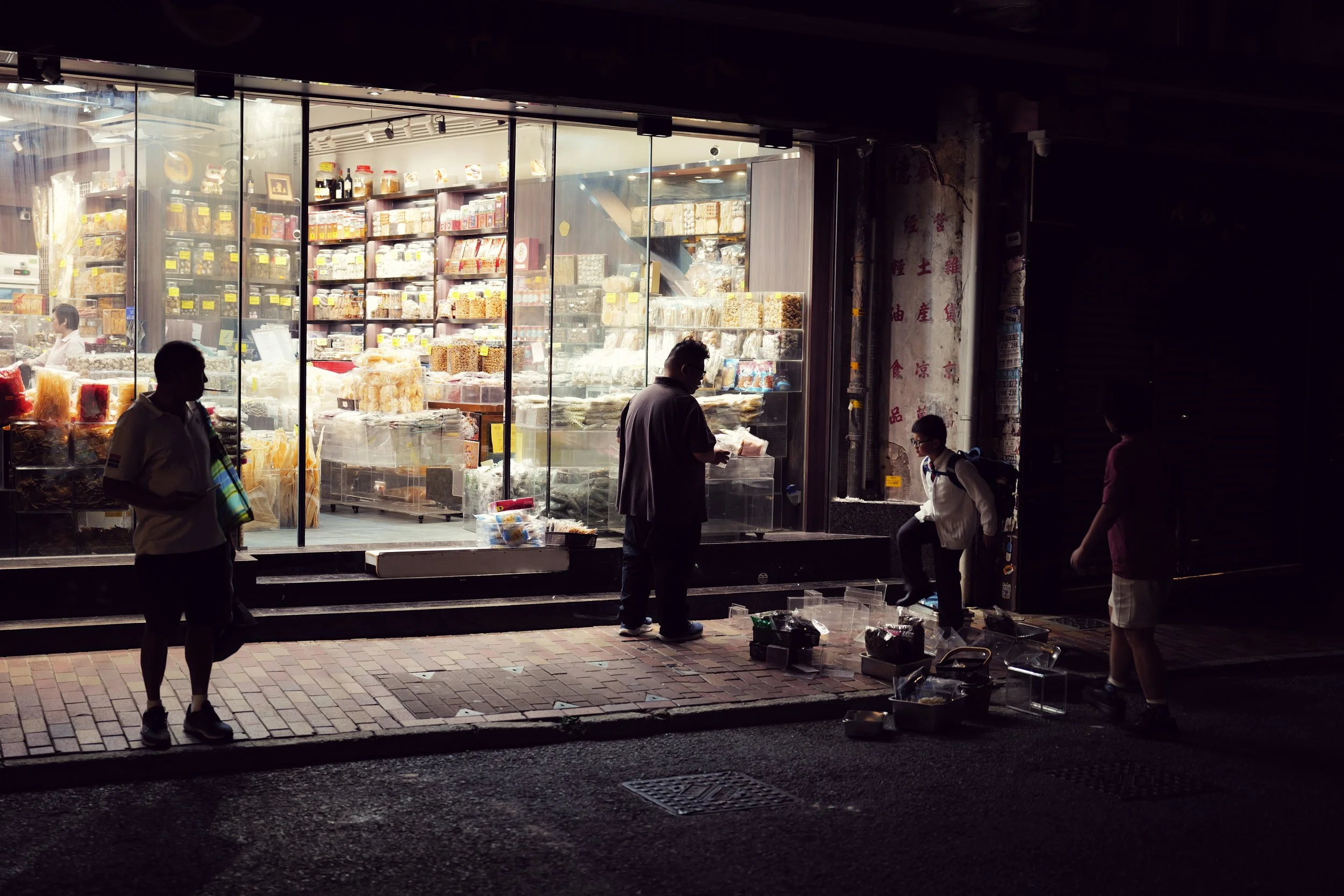 Night view of a store with glass windows displaying various packaged goods. Several people are outside, some looking at the products and some walking by, with street lights illuminating the sidewalk.