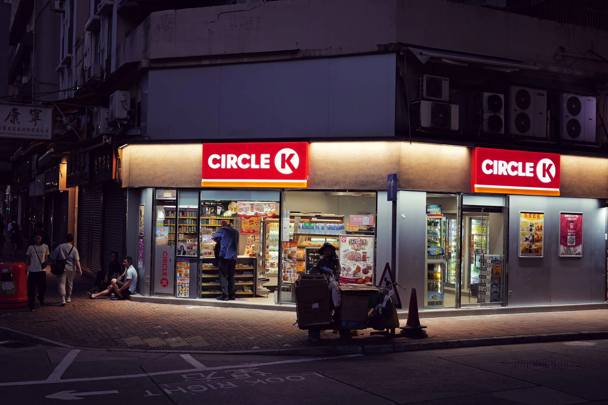 A street view of a convenience store called Circle K with illuminated signs, people walking and standing outside, and vending machines visible in the foreground, during evening or nighttime.