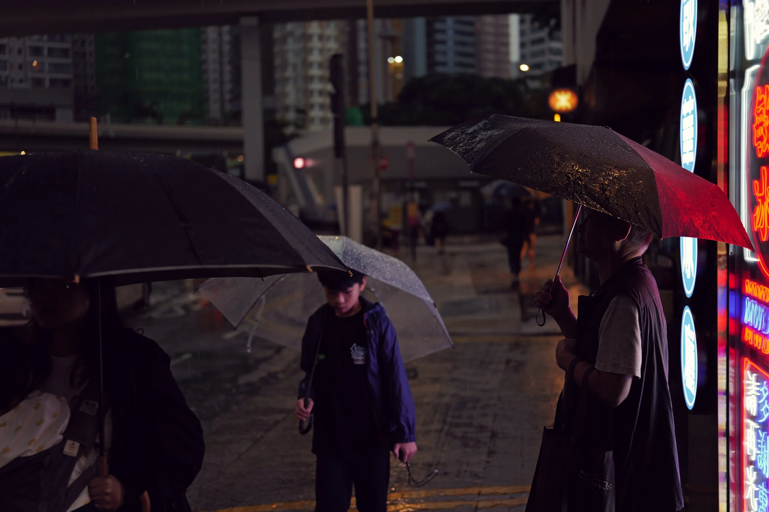 People walking outdoors at night with umbrellas in a rainy city, illuminated by neon signs.