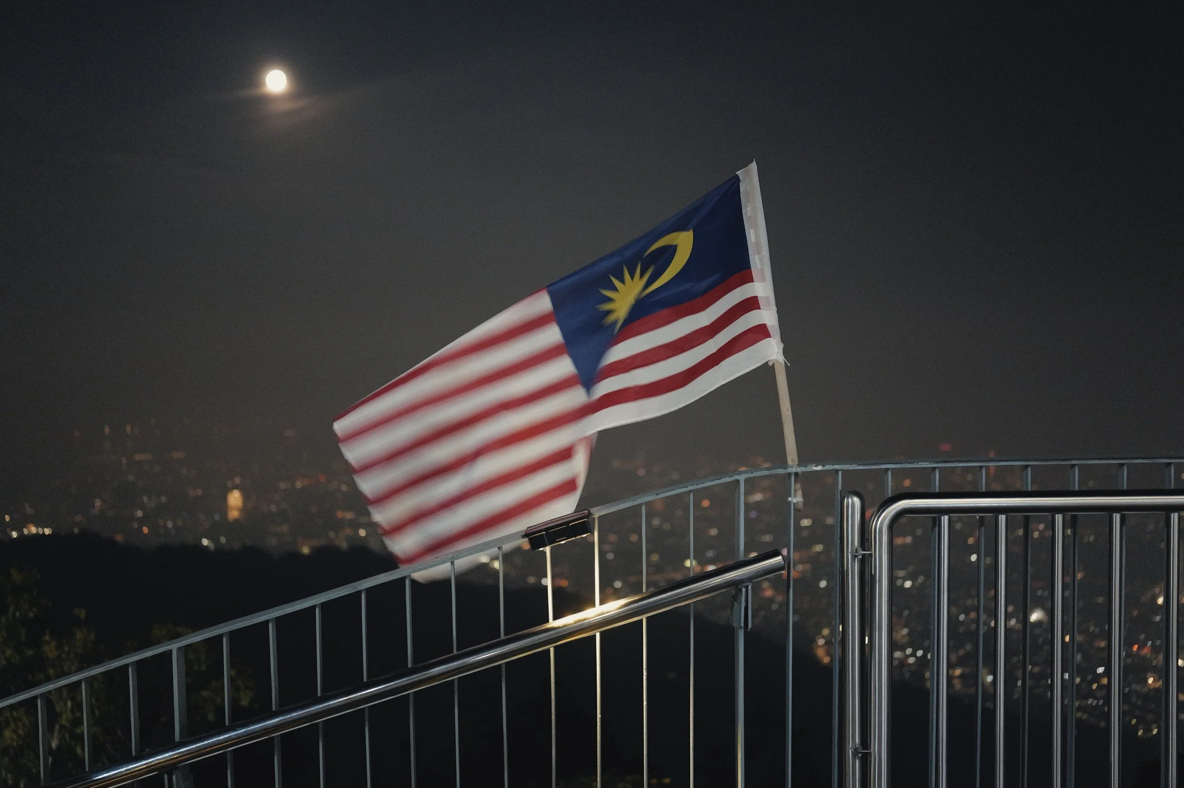 A Malaysian flag spreads above a balcony railing with city lights and the moon beyond.