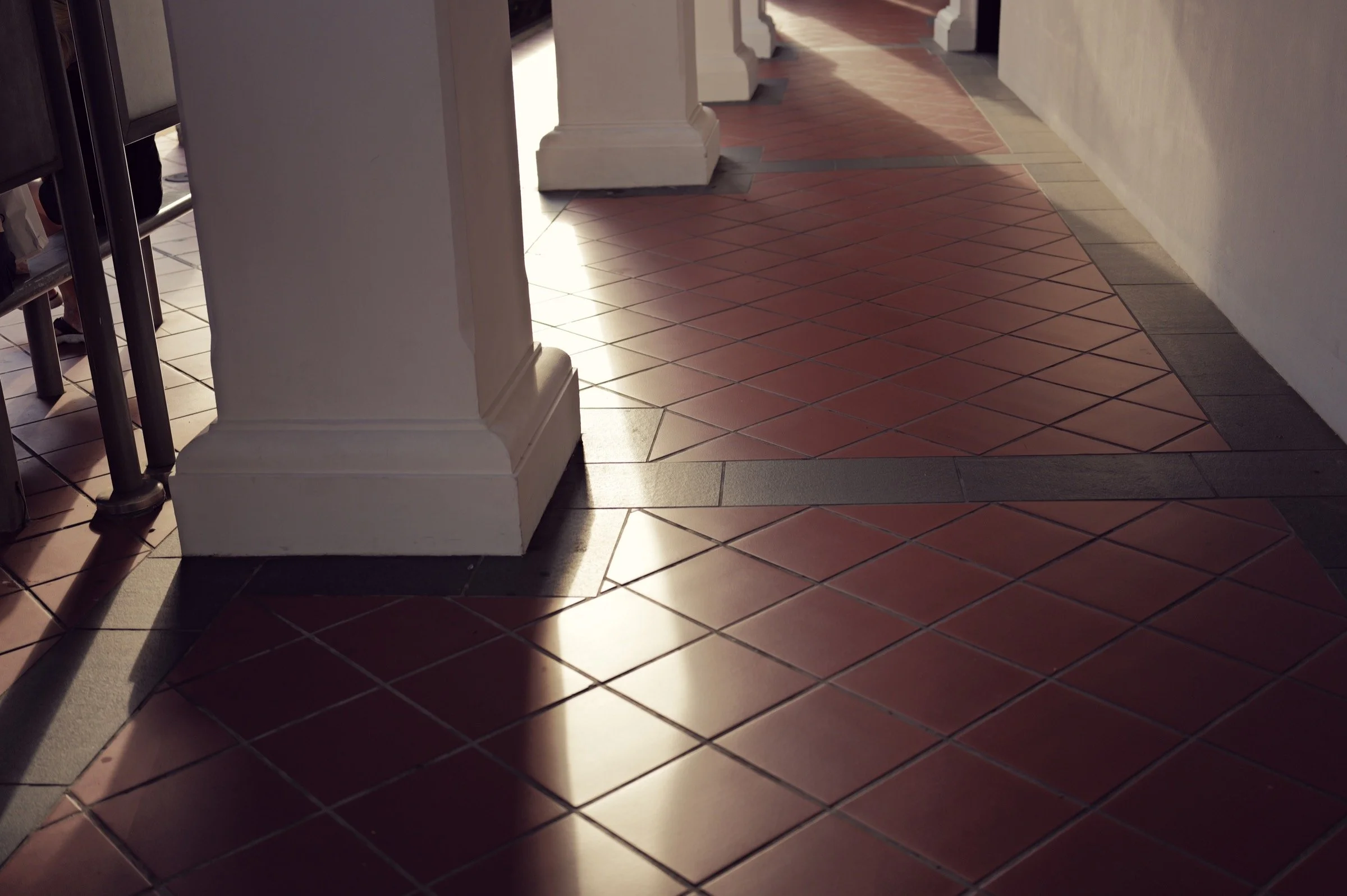 Sunlight cuts across red and dark floor tiles between white columns in a covered walkway.