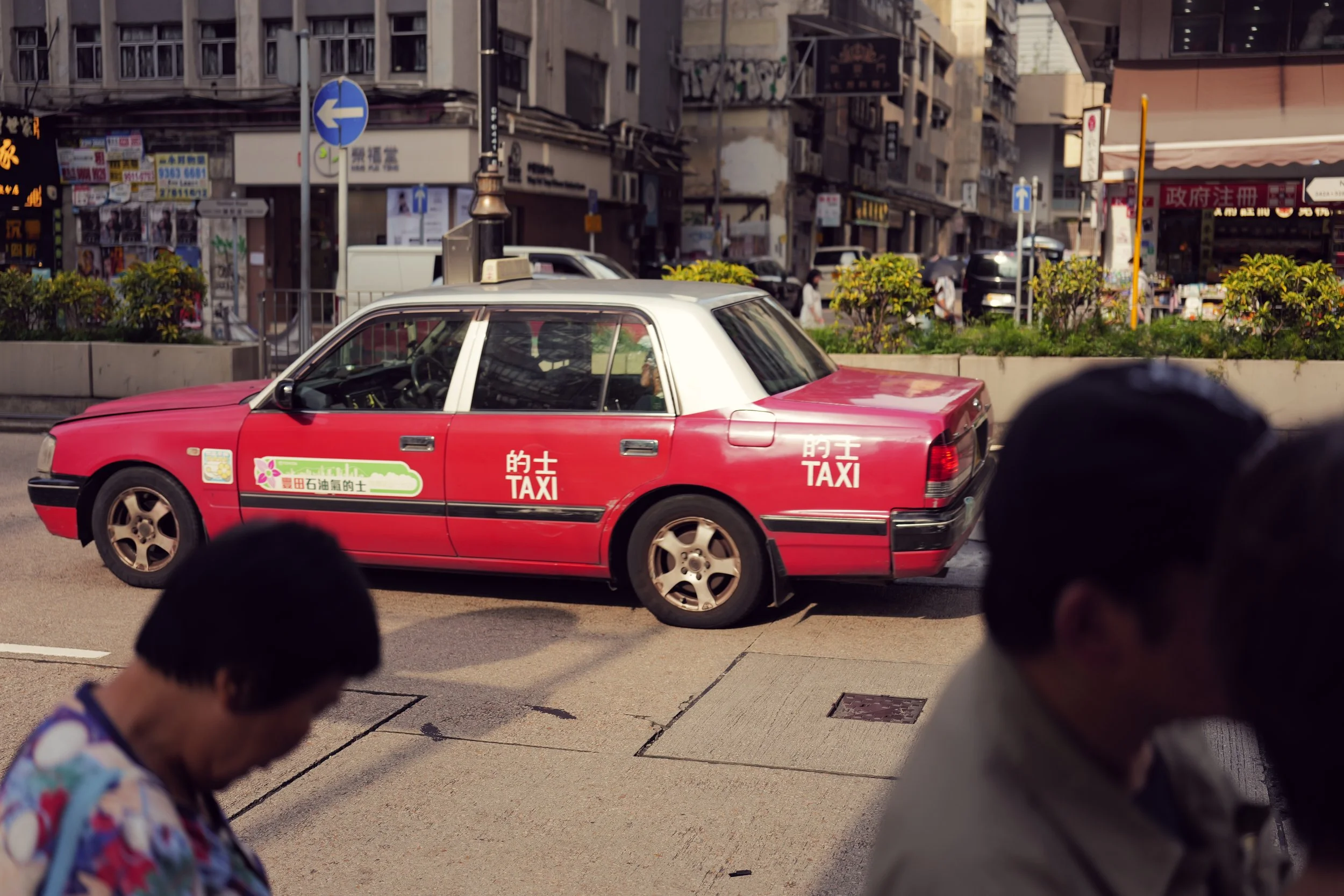 A red and white taxi cab driving on a city street with buildings and signage in the background. Several pedestrians are walking on the sidewalk, and there are street signs for traffic directions.