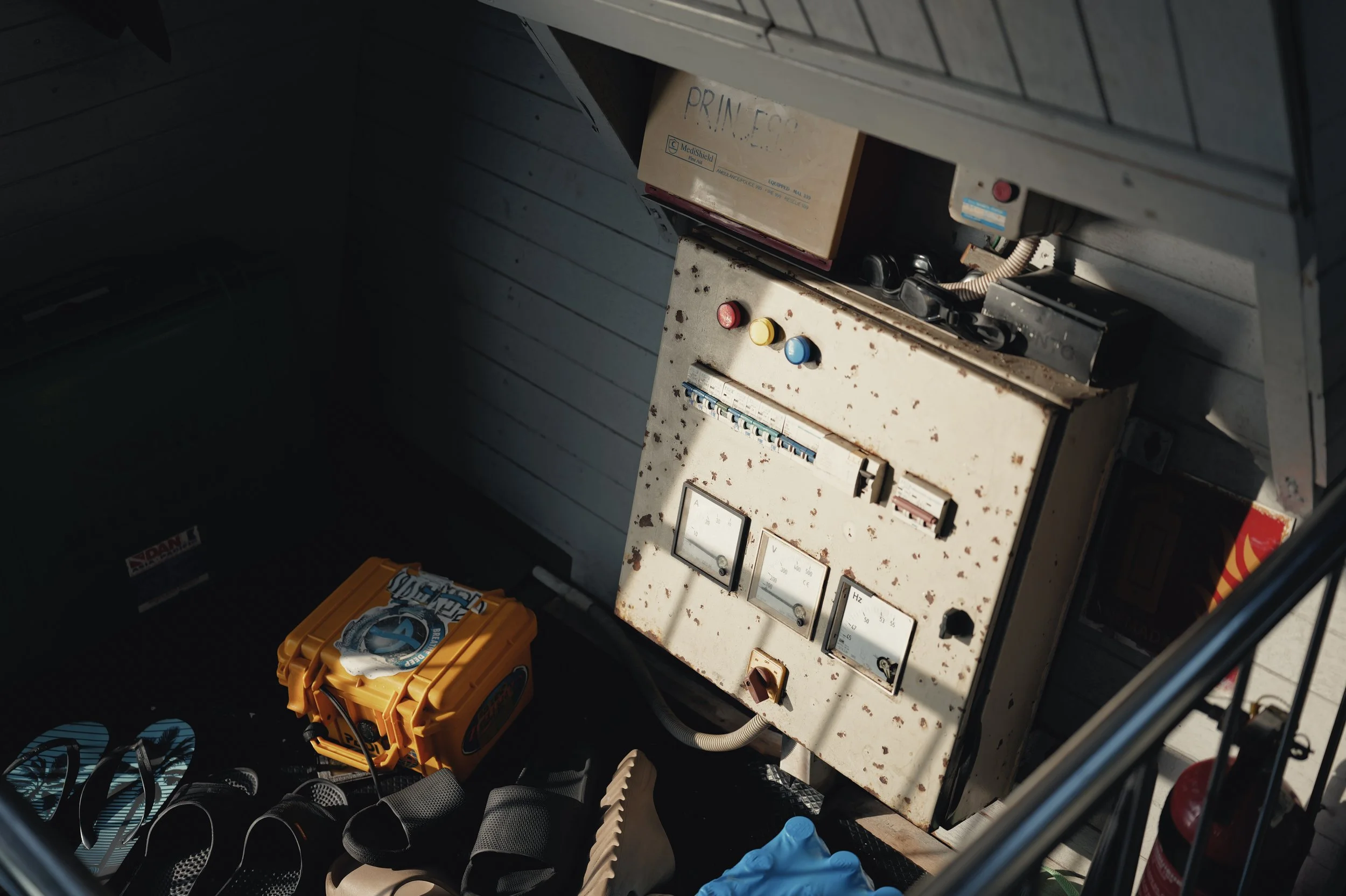 An industrial control box with rust and dirt, featuring three colored buttons (red, yellow, blue) and gauges. A cardboard box labeled 'PRIMECO' is on top of it. To the side, there are black sandals and a yellow toolbox with a logo, along with a pair 