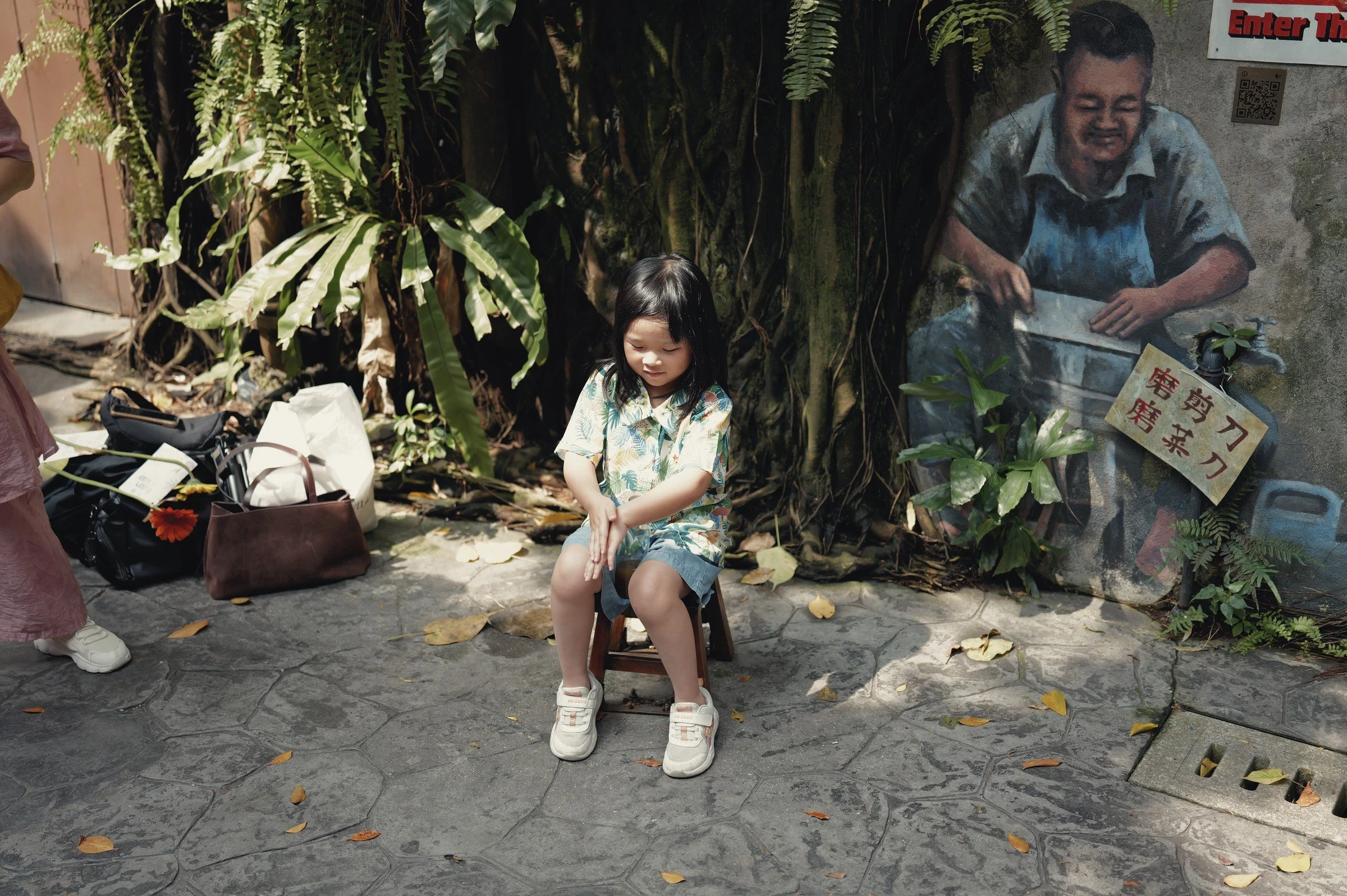 A visitor sits beneath hanging greenery beside a painted street mural.