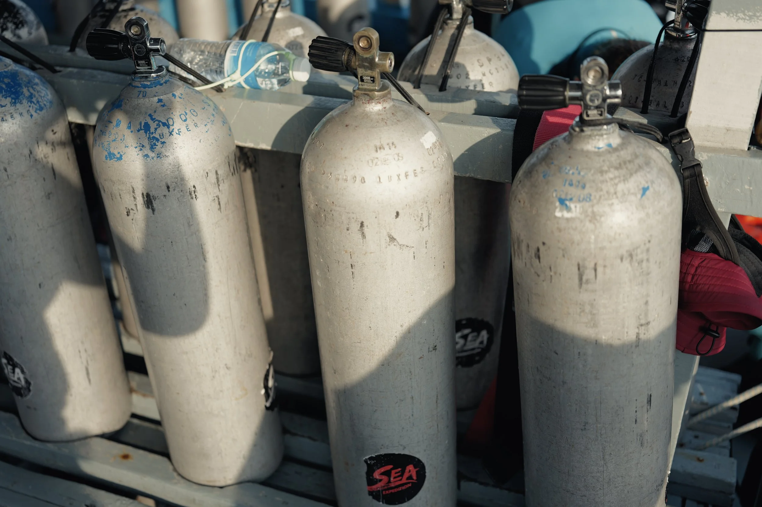 A row of white scuba diving tanks with black valves and hoses, some with blue paint markings, secured on a surface. A plastic water bottle is resting on top of some tanks, and part of a red bag with a black strap is visible.