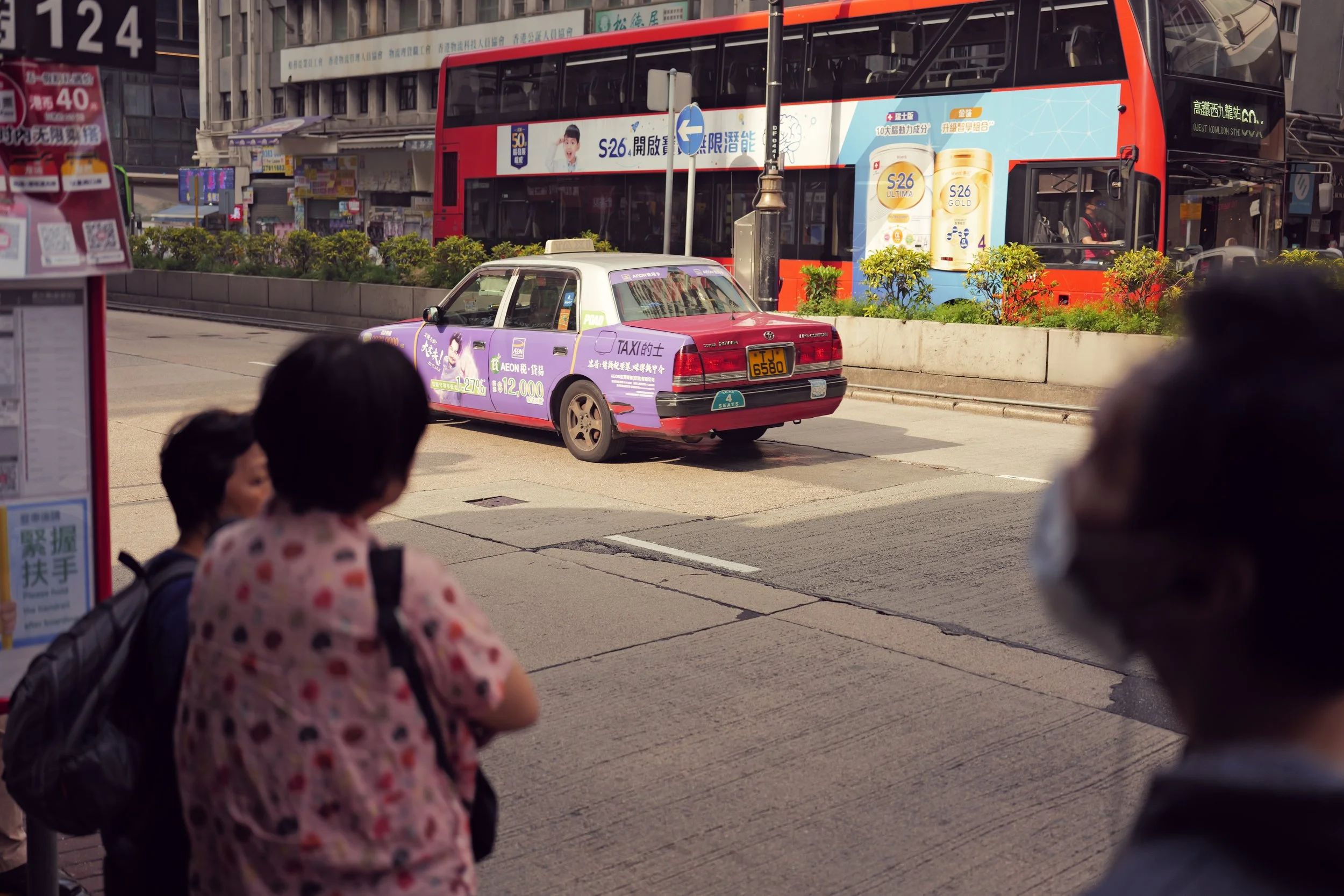 A street scene showing a purple taxi cab with advertisements on it, in front of a double-decker bus with advertisements on it, and some pedestrians standing at a bus stop.