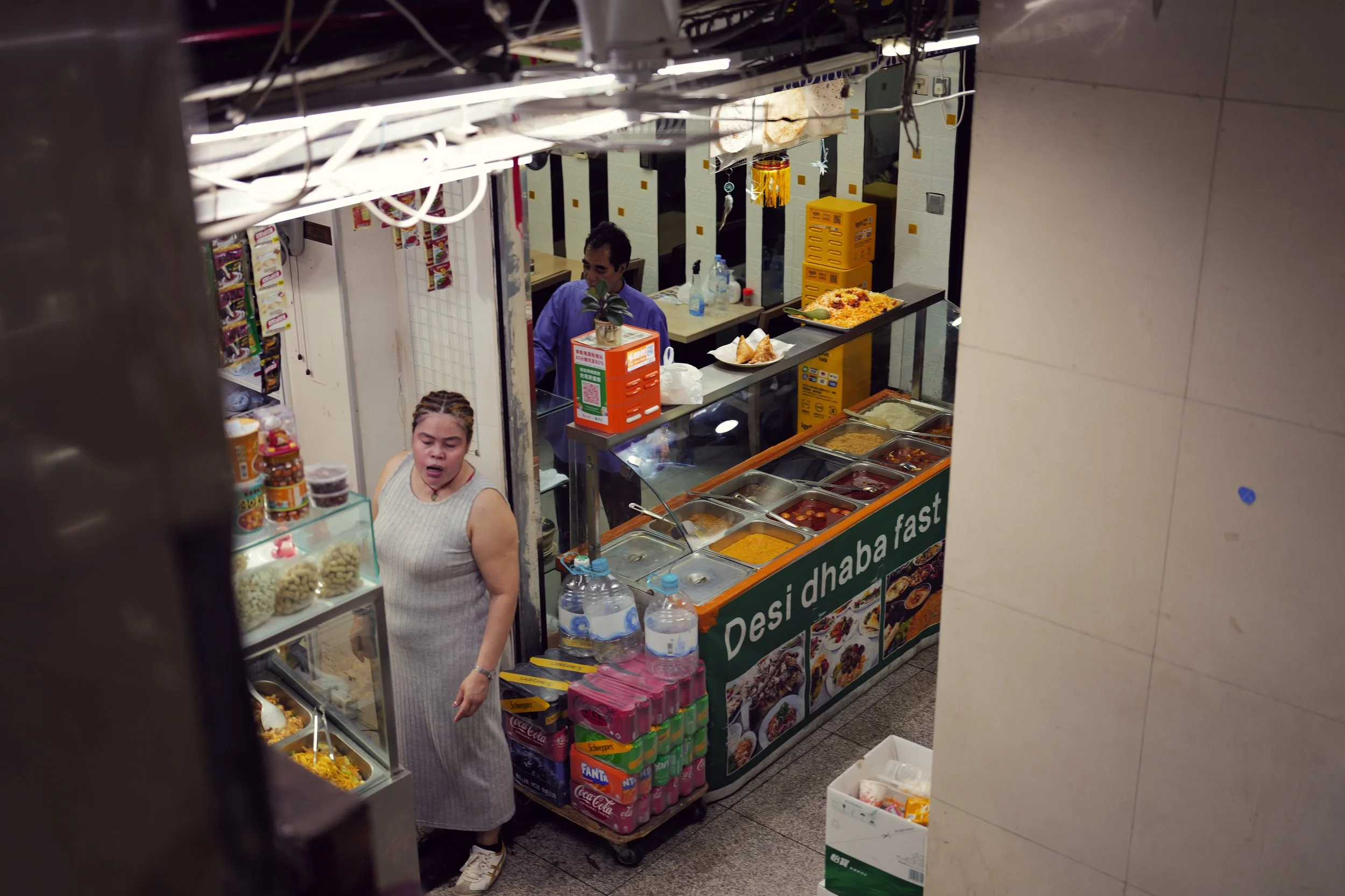 A small food stand with various dishes and drinks, and two people serving or ordering. The stand has containers of food, bottles of drinks, and a sign for desi dishes, located indoors near tiled walls.