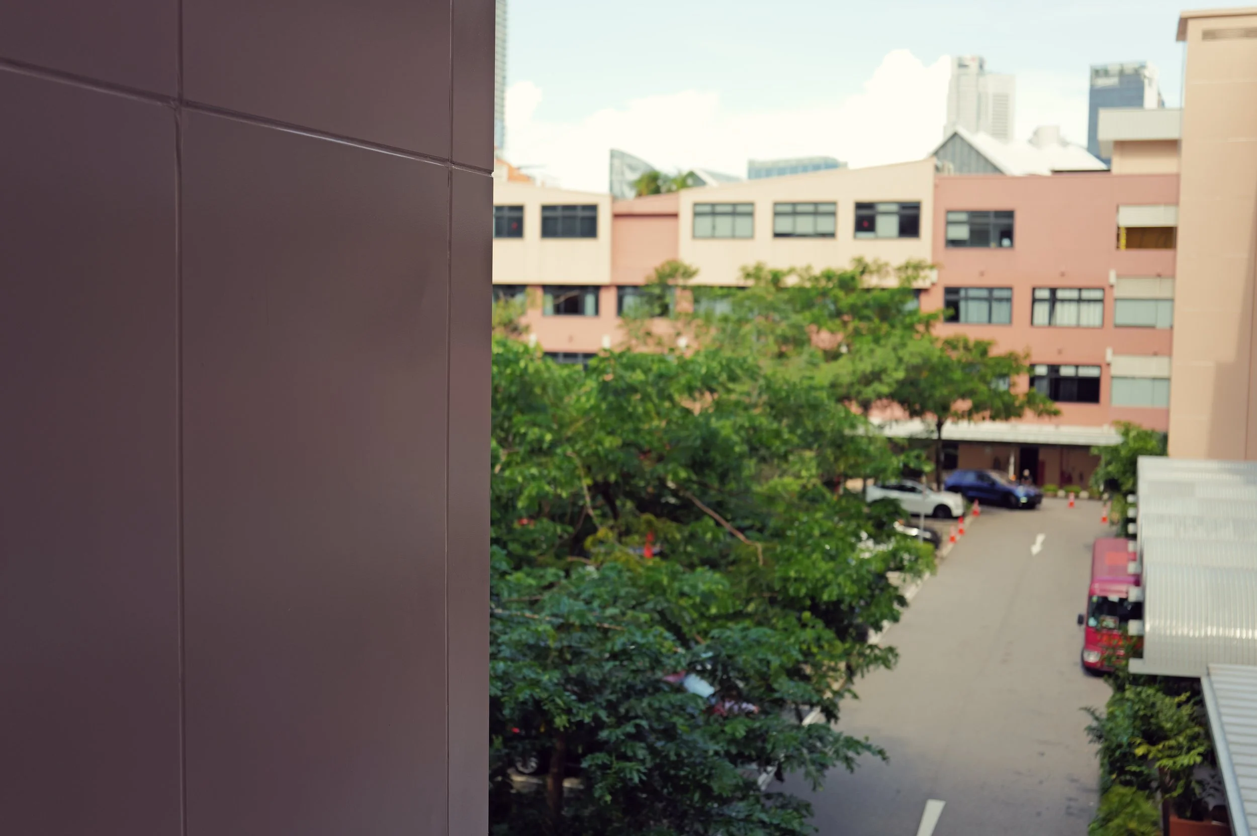 View from a building showing a parking lot with cars, a green tree, and an exterior of a pinkish beige building with many windows in an urban area with tall buildings in the background.