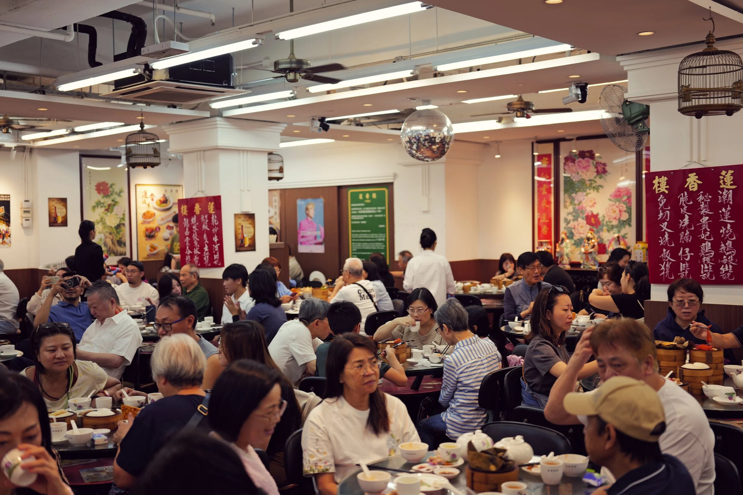 A large indoor restaurant filled with many seated people enjoying a meal. The restaurant features bright lighting, decorative wall art with floral themes, hanging birdcages, and a disco ball on the ceiling.
