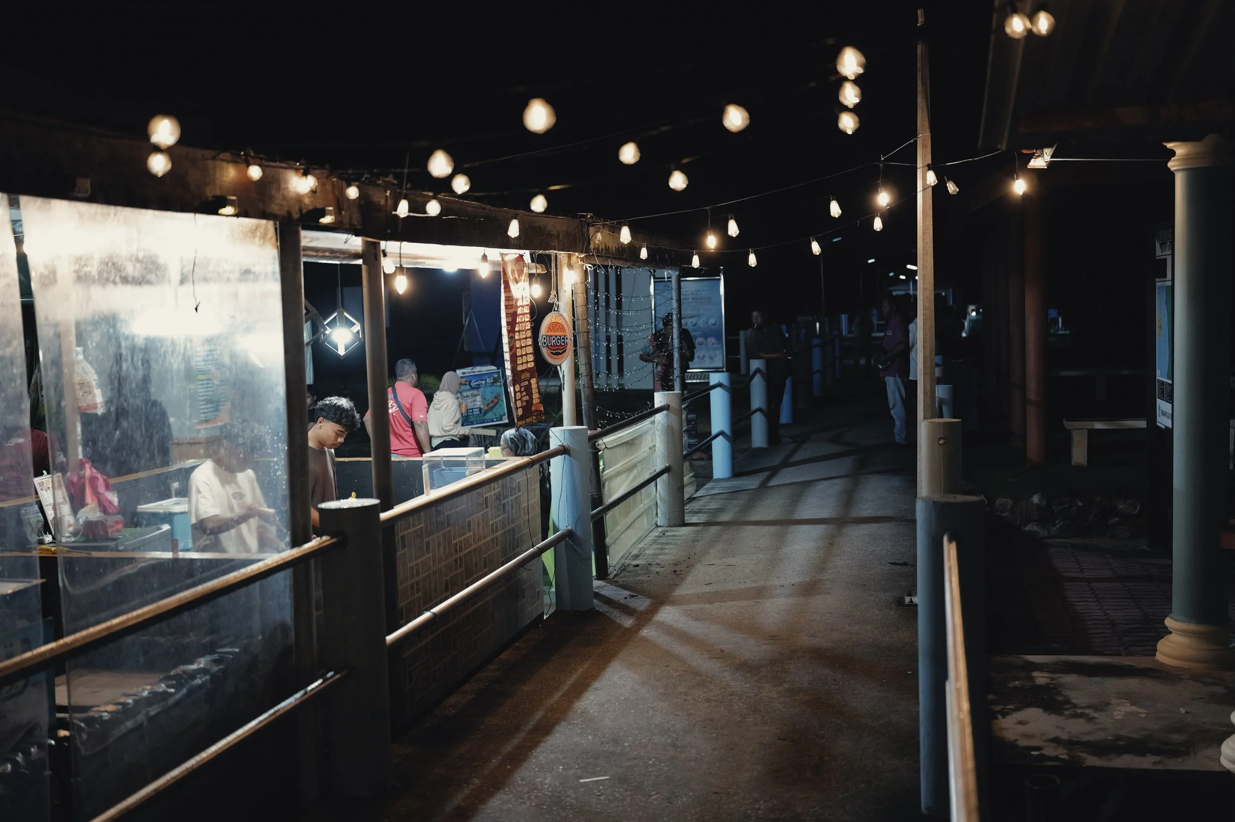 Night scene at a food stall with string lights overhead, people standing in line, and a illuminated burger sign in the background.
