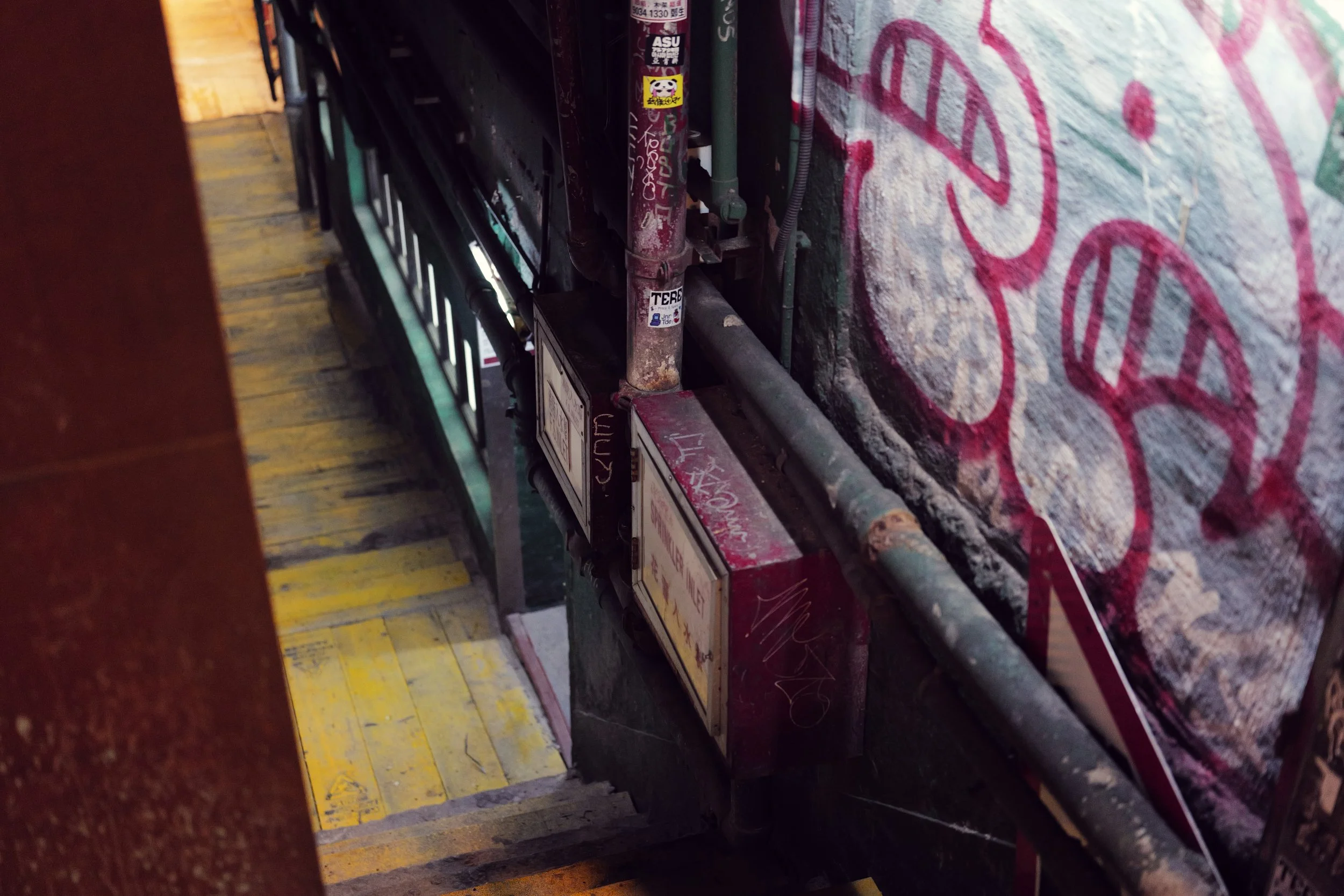 A narrow indoor space with a yellow-stained wooden floor and a wall with graffiti. The perspective is from a low angle looking along a surface with various overlapping objects, including stickers and a pipe.