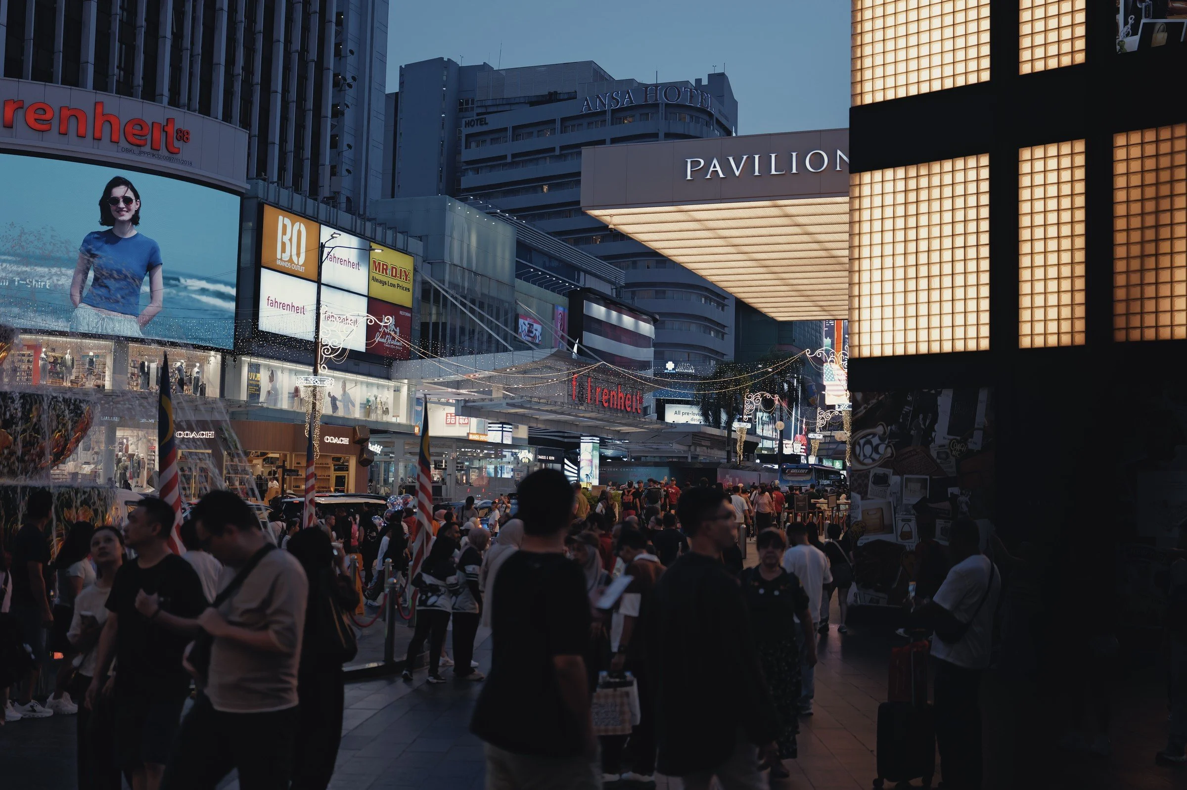 Crowds pass illuminated signs and shopping buildings at dusk.