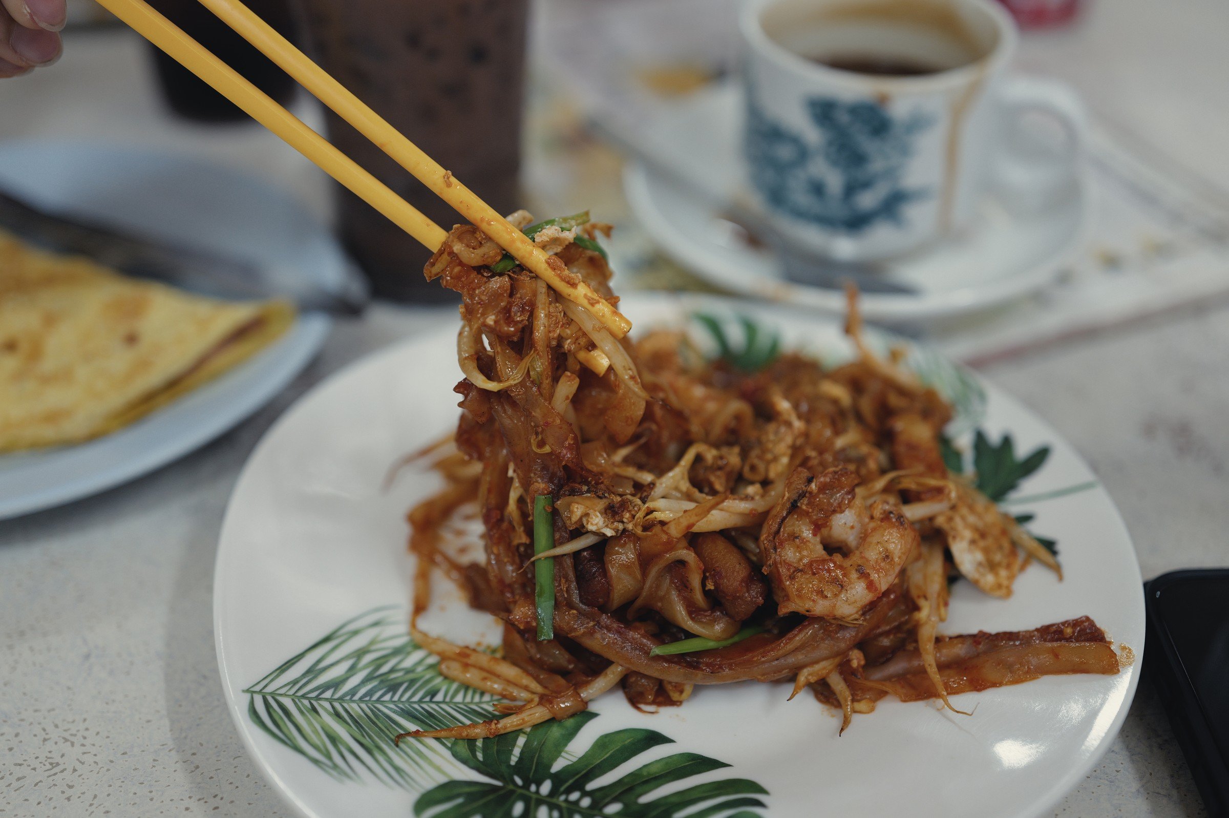 Chopsticks lift fried noodles above a patterned plate beside a cup of coffee.