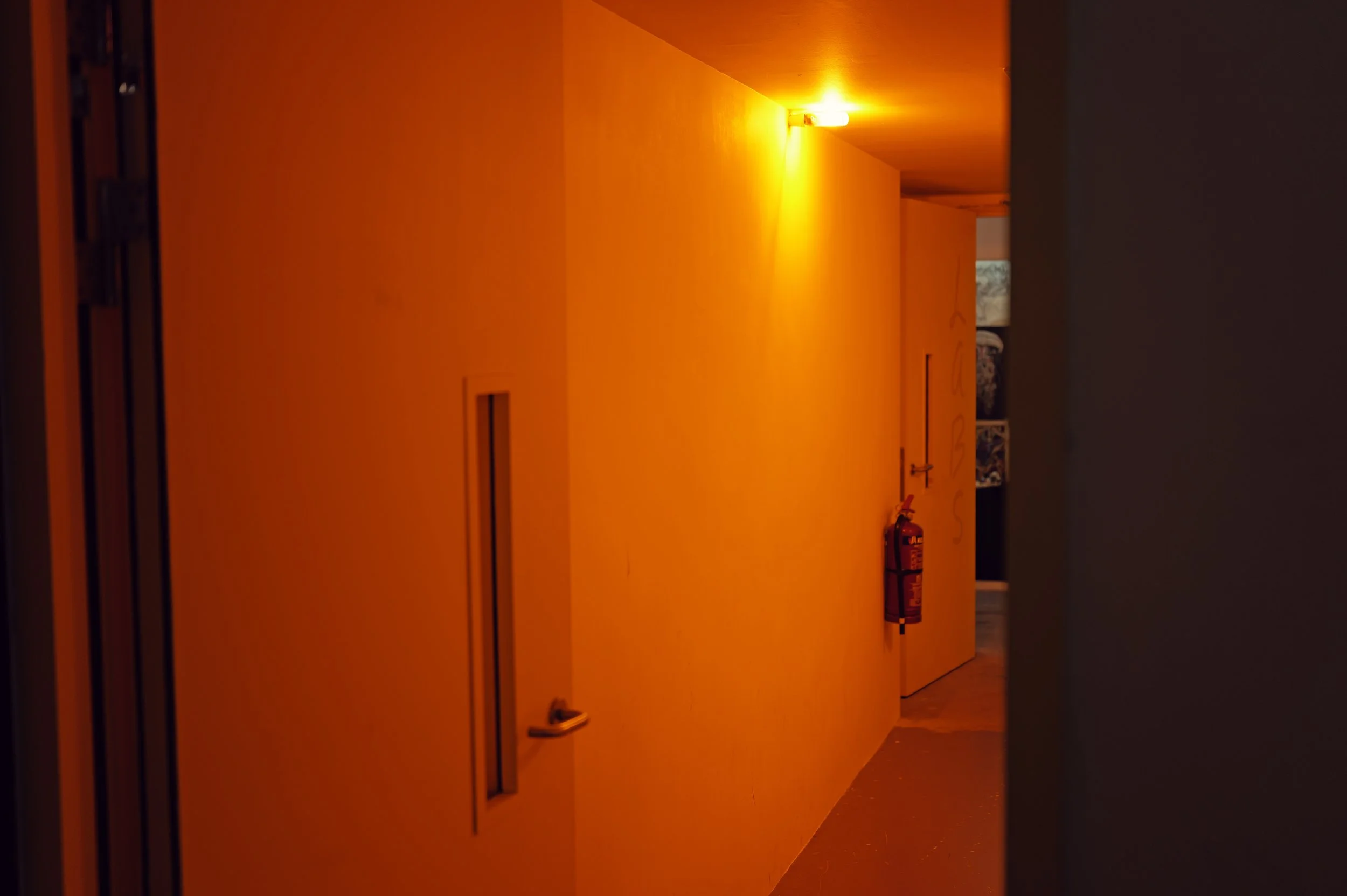 A hallway with orange walls and ceiling, illuminated by a yellowish light near the ceiling. There is a fire extinguisher mounted on the wall near the lower right corner. A door is visible at the end of the hallway, and a window can be seen at the far