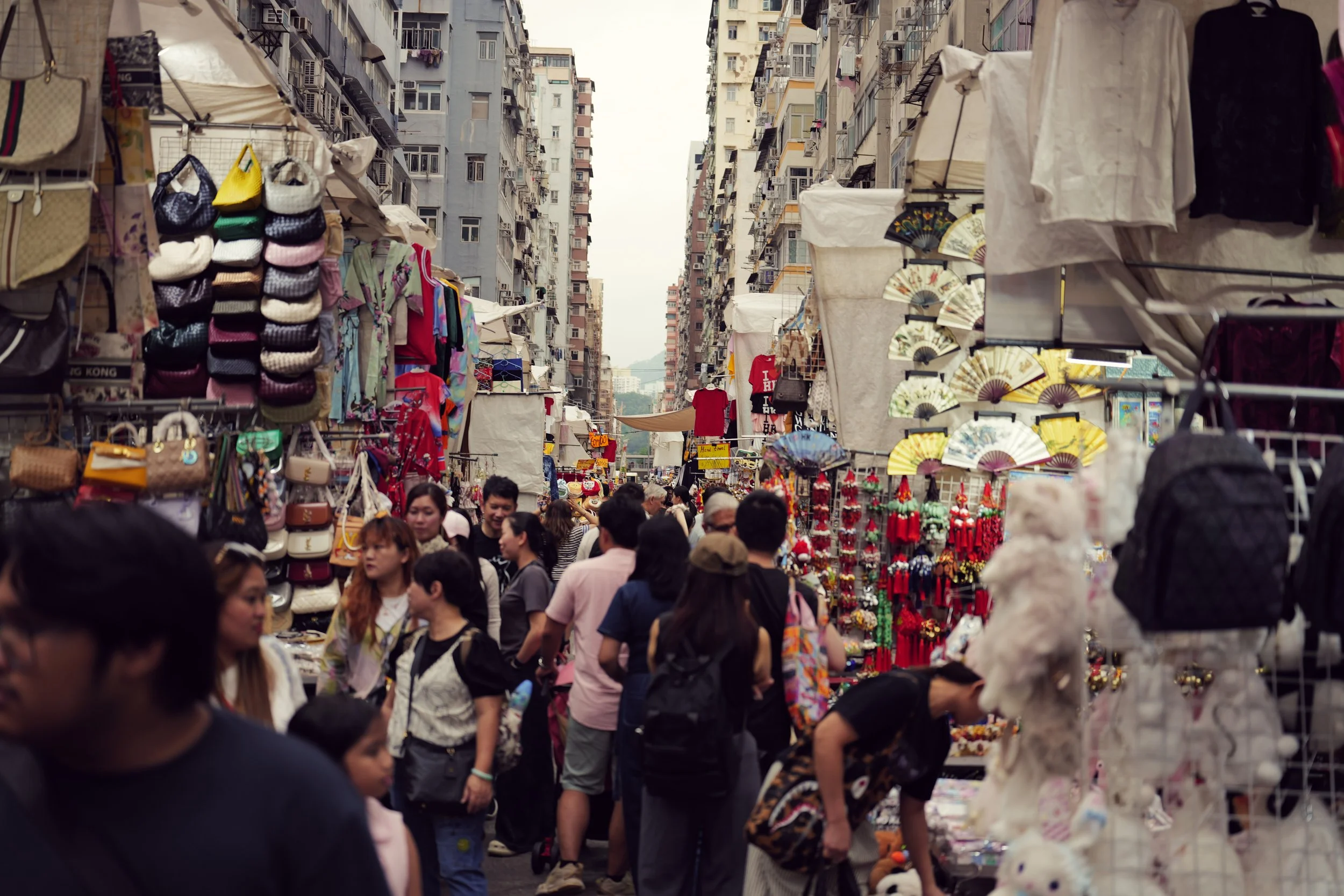 A crowded outdoor marketplace with stalls selling bags, fans, clothing, and accessories. Tall residential buildings are visible in the background.